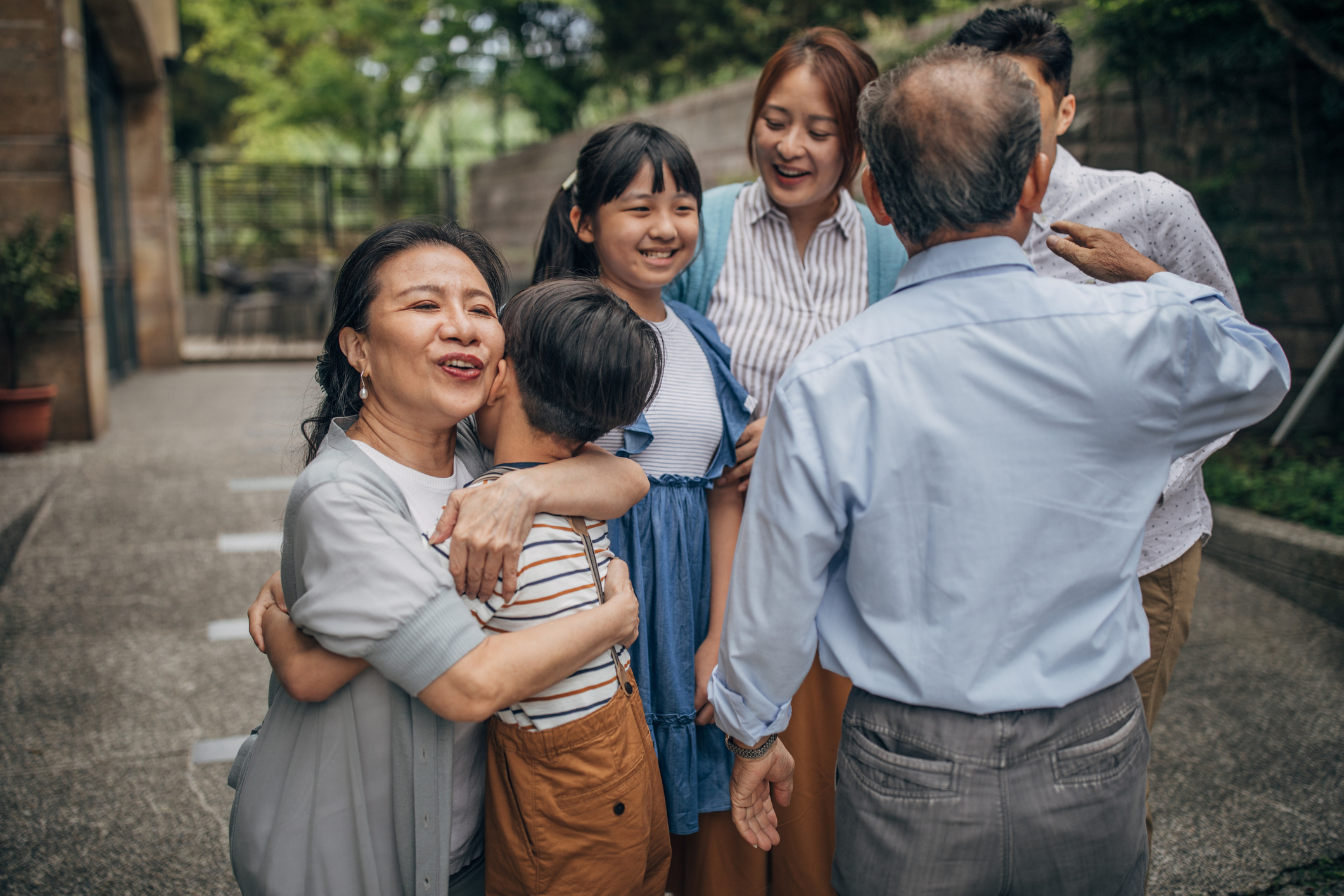 Group of six people, including grandparents, parents, and children, in an outdoor setting, smiling and hugging each other in a warm family moment