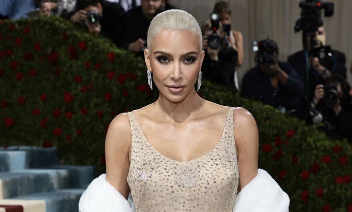 Kim Kardashian poses on the red carpet in a sparkling, sleeveless gown with a plunging neckline, accessorized with drop earrings and a white shawl draped over her arms