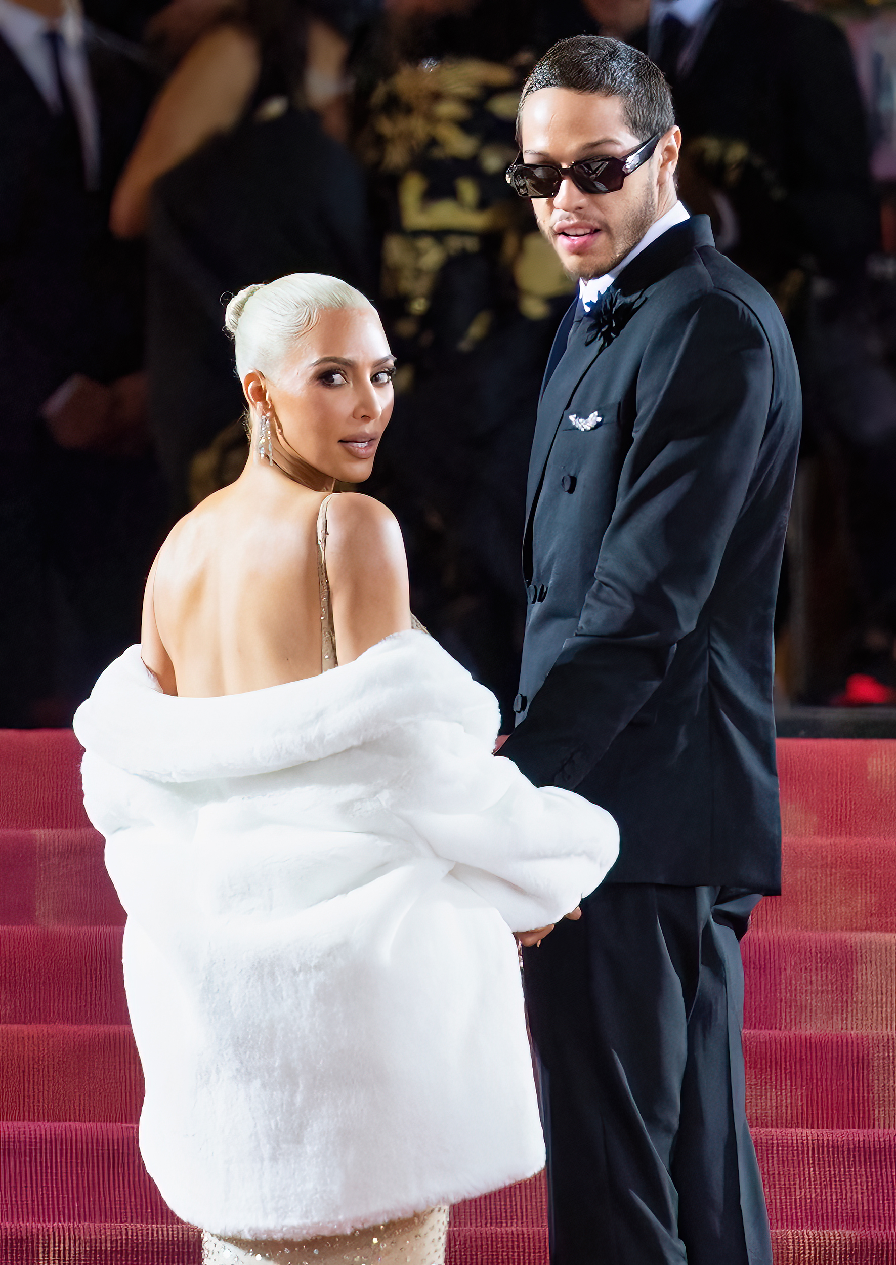 Kim Kardashian and Pete Davidson pose together on a red carpet. Kim wears an elegant strapless gown with a fluffy shawl, while Pete wears a stylish suit with sunglasses
