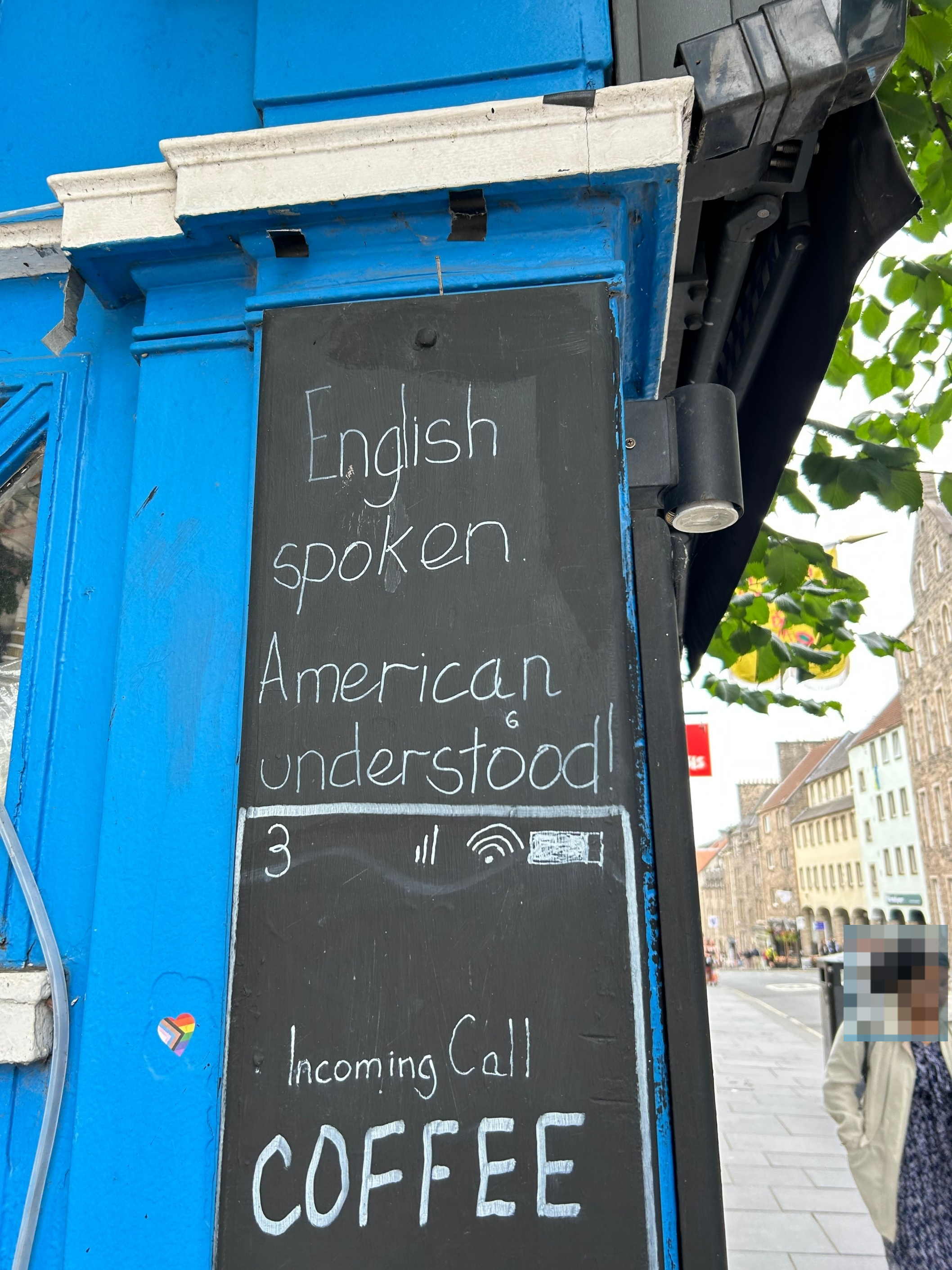 Sign on a building reads: "English spoken, American understood. Incoming Call: COFFEE." People walking nearby