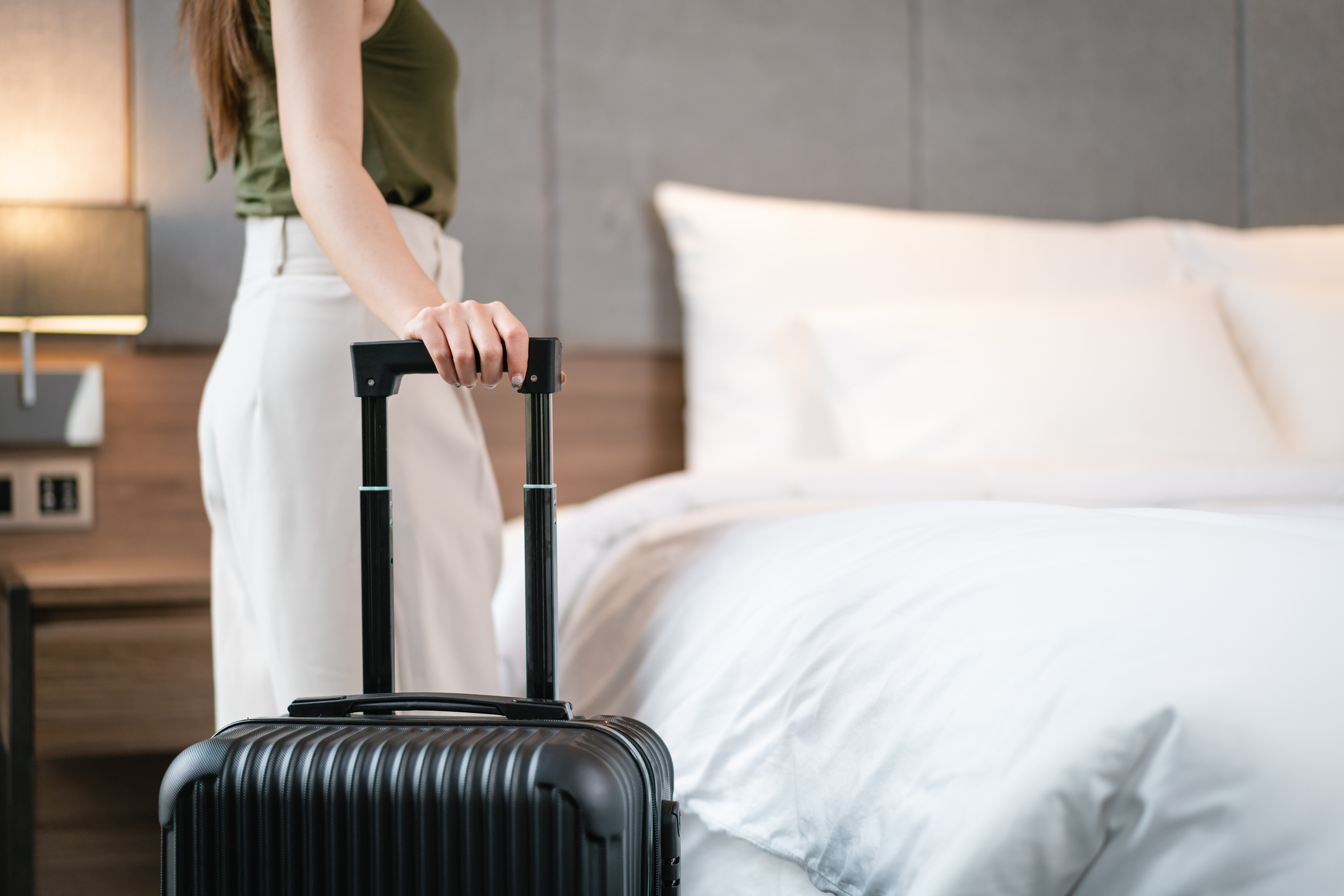 A person standing beside a hotel bed, holding the handle of a black suitcase. The individual is dressed in casual attire, ready for travel