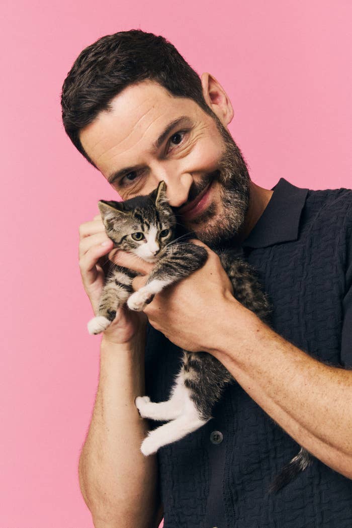 Tom Ellis smiles, holding a kitten close to his face against a pink background. He is wearing a textured, short-sleeved shirt
