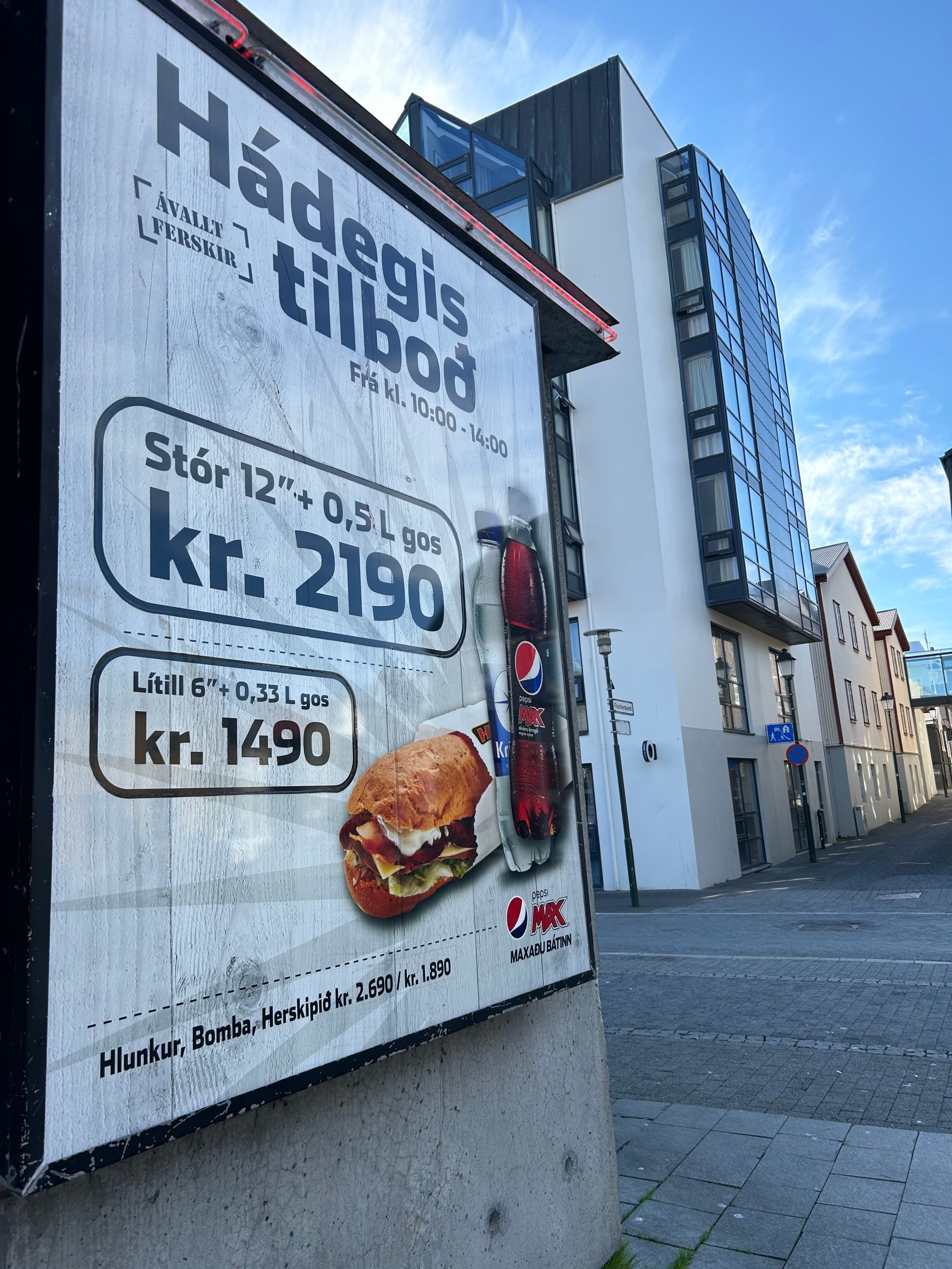 An outdoor displays a lunchtime offer for a sandwich with a drink for kr. 2190 and a smaller meal for kr. 1490. A city street with buildings is in the background