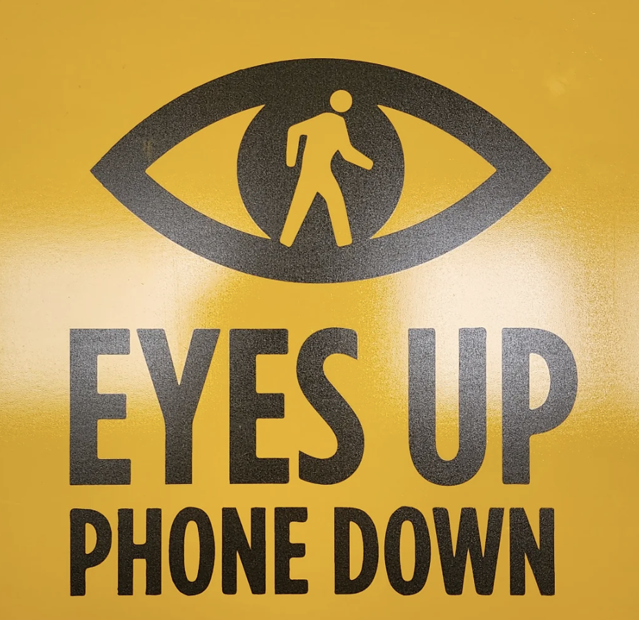 A sign with an eye symbol and a walking person inside the eye. The text reads &quot;EYES UP PHONE DOWN.&quot;