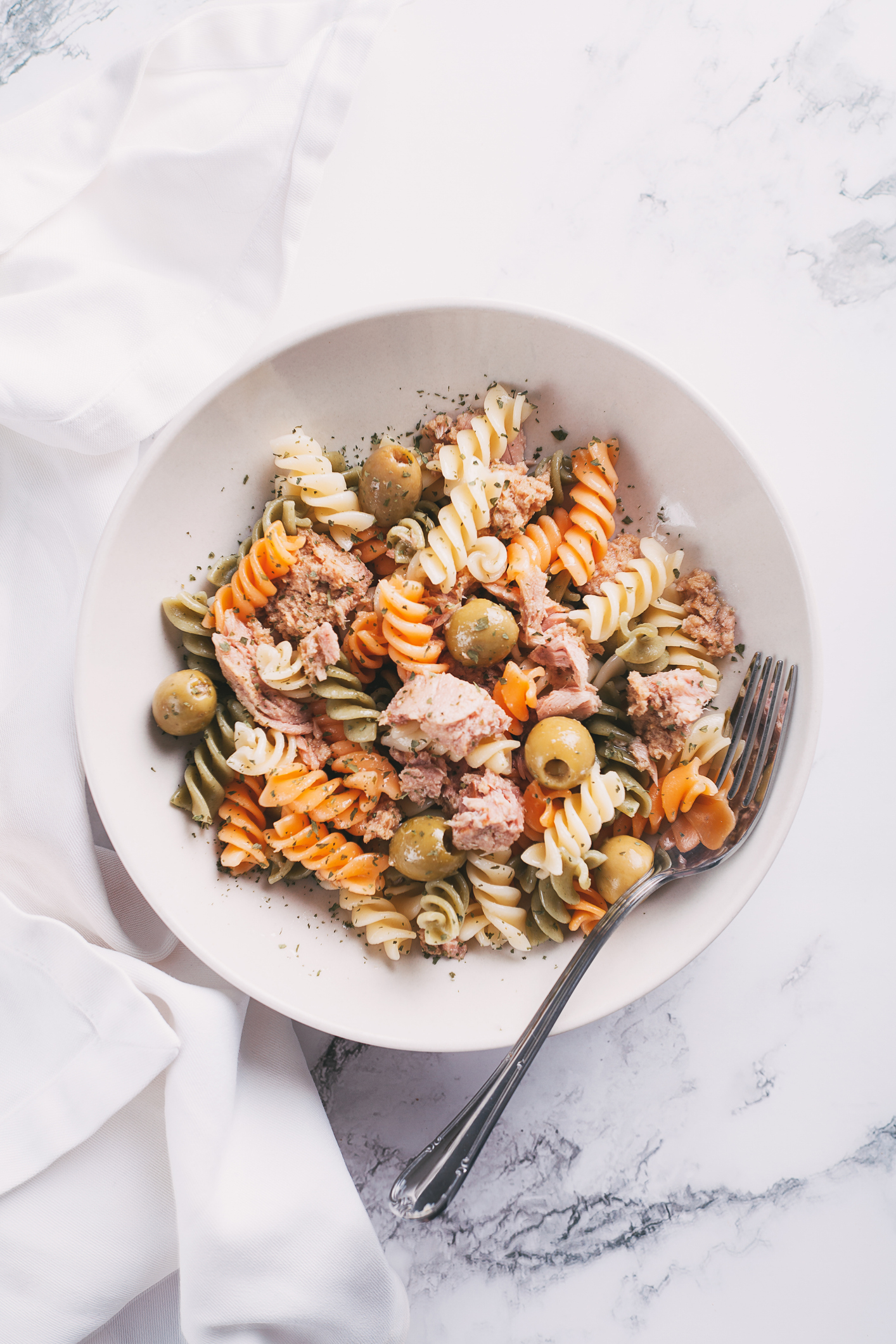 A bowl of tri-color rotini pasta mixed with tuna chunks and green olives, accompanied by a fork on the side