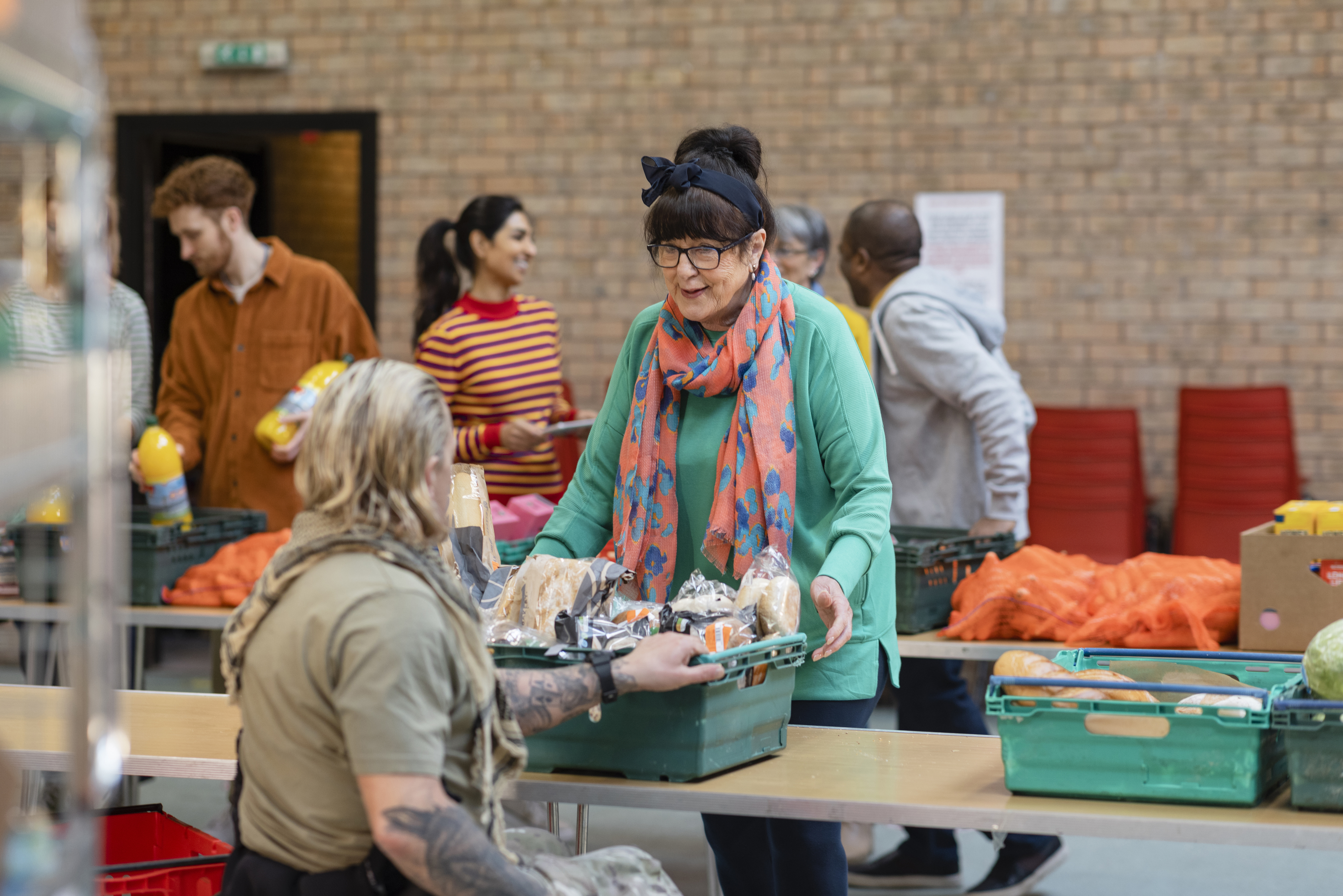 People volunteer, distributing food at a community center. One woman accepts a crate of food from another volunteer with tattoos on their arm
