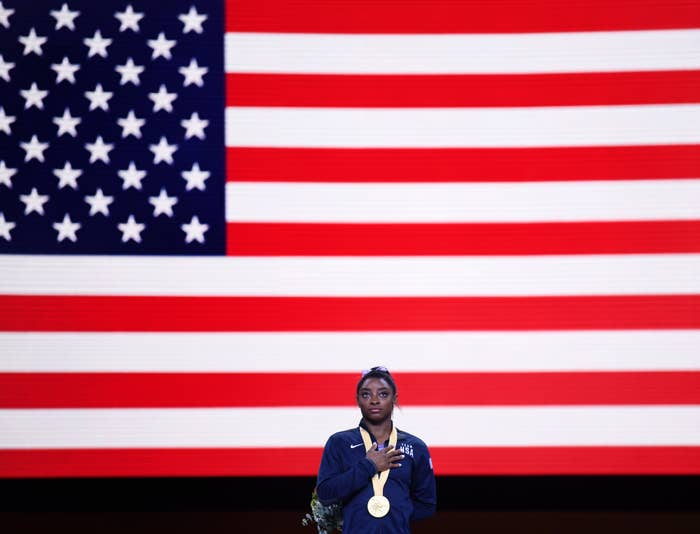 Simone Biles stands with her hand over her heart, wearing a medal in front of a large American flag