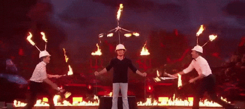 Simon Cowell stands between two people juggling flaming items, with fire in the background on a TV set