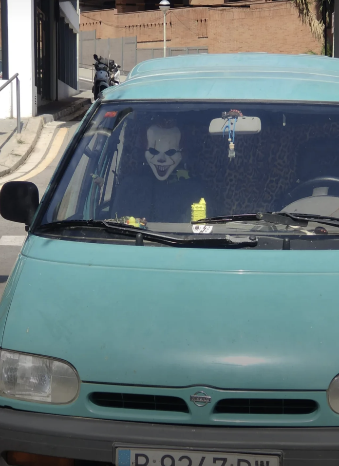 A scary clown mask is placed behind the wheel of a parked van, giving the appearance that it's driving