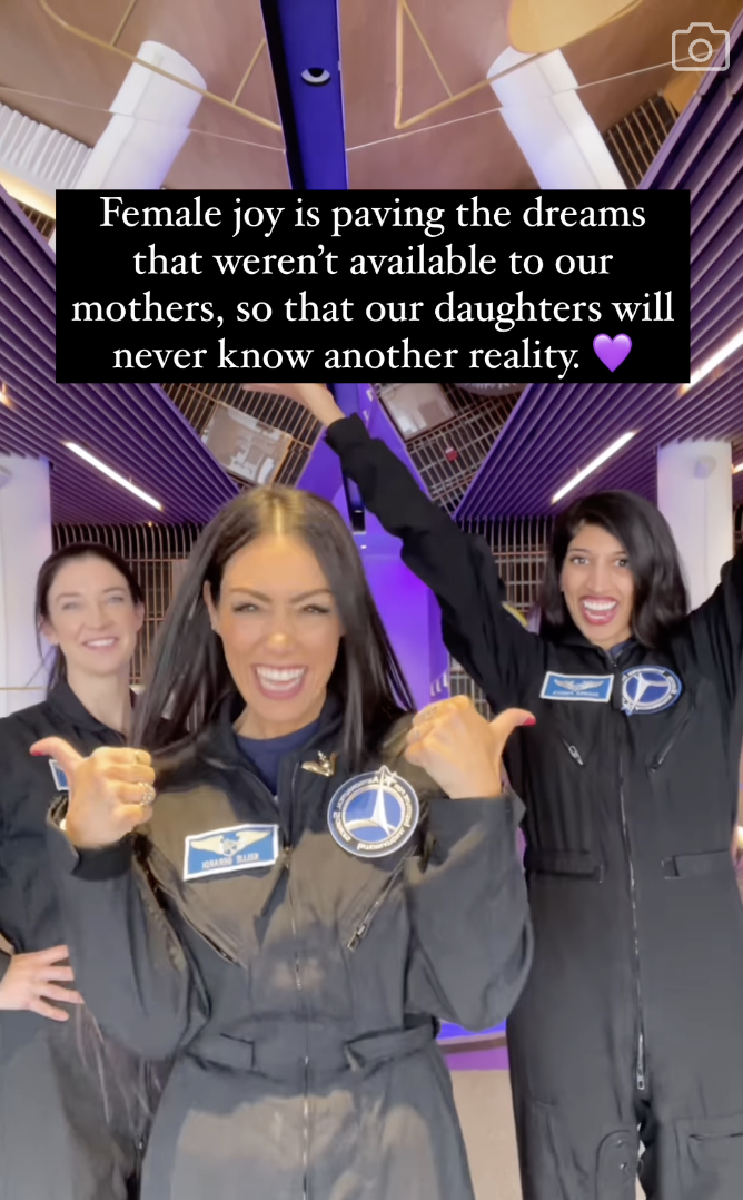 Three women in astronaut uniforms celebrate under a lit ceiling with text above them stating the joy of pursuing dreams for future generations
