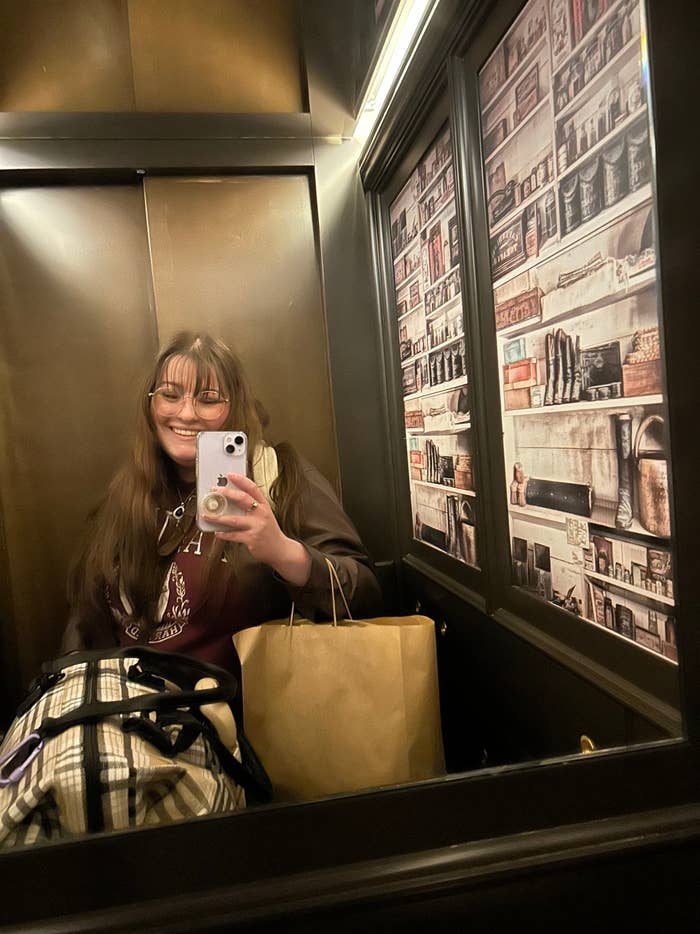 Kristen with long hair and glasses takes a selfie in an elevator with walls decorated to look like bookshelves. She holds a shopping bag and a plaid-patterned bag