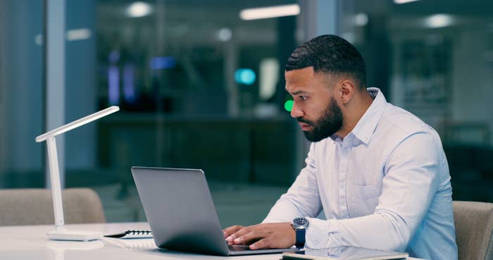 A person at a desk concentrates while working on a laptop in a modern, dimly-lit office. They are dressed in a button-down shirt