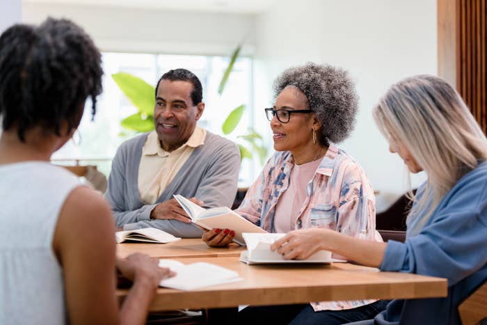 A group of four people sit at a table, smiling and discussing books. They appear in a brightly lit room. Names of the people are not provided