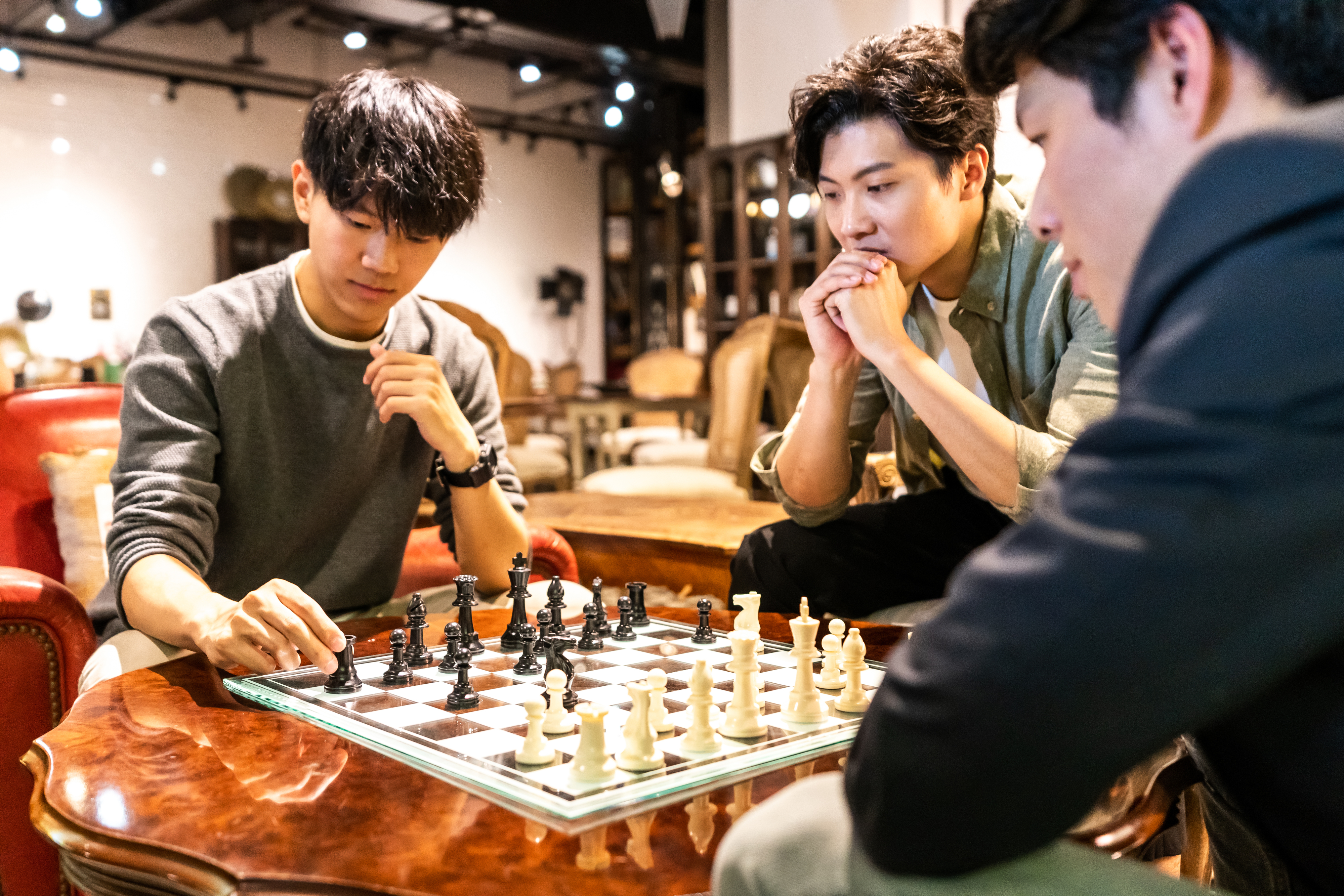 Three men focused on an intense game of chess at a cozy, well-lit room with wooden furniture. Their expressions reflect deep concentration