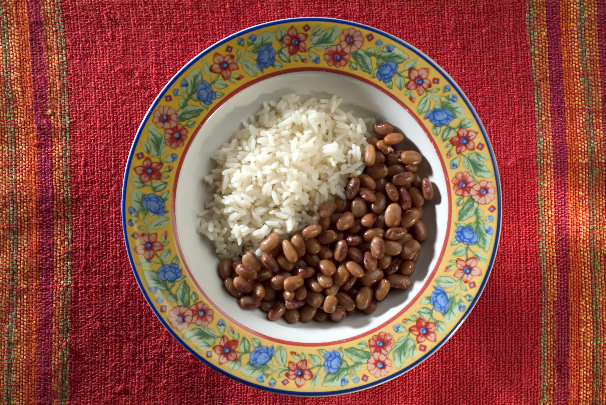 A bowl of white rice and beans, served on a decorative plate with a floral border, placed on a red woven tablecloth