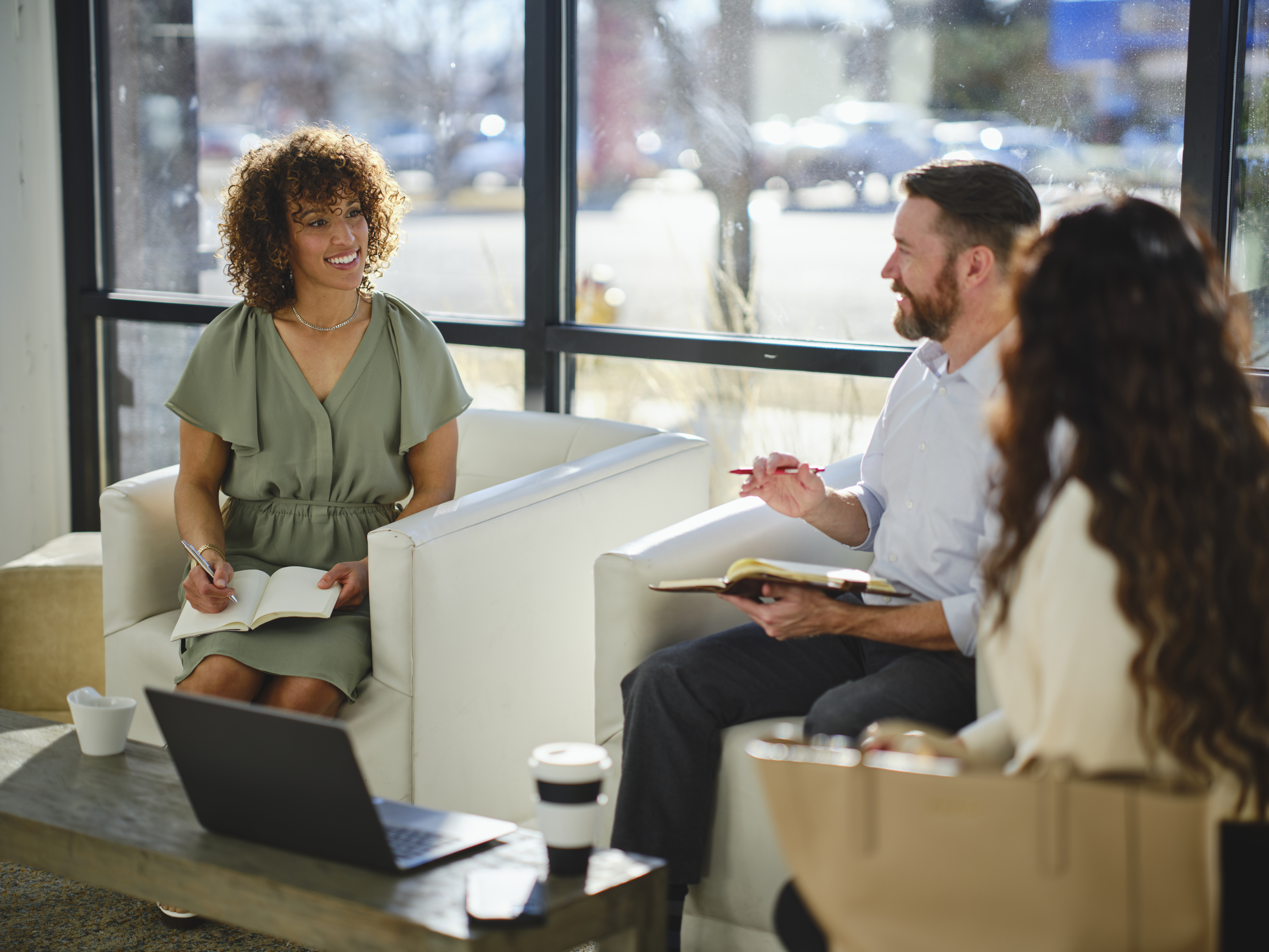Three people having a friendly meeting in a bright room. One woman is holding a notebook, another woman is seated, and a man is speaking to them