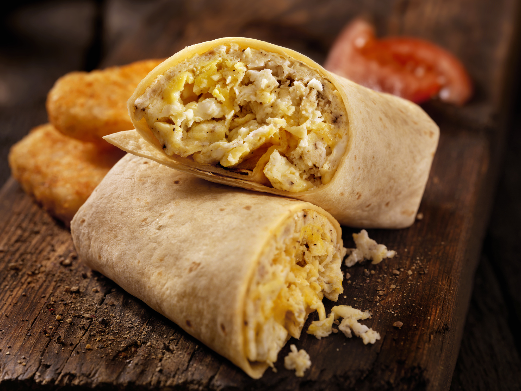 A close-up of a breakfast burrito cut in half, filled with scrambled eggs and cheese, placed on a wooden surface next to hash brown patties and a slice of tomato