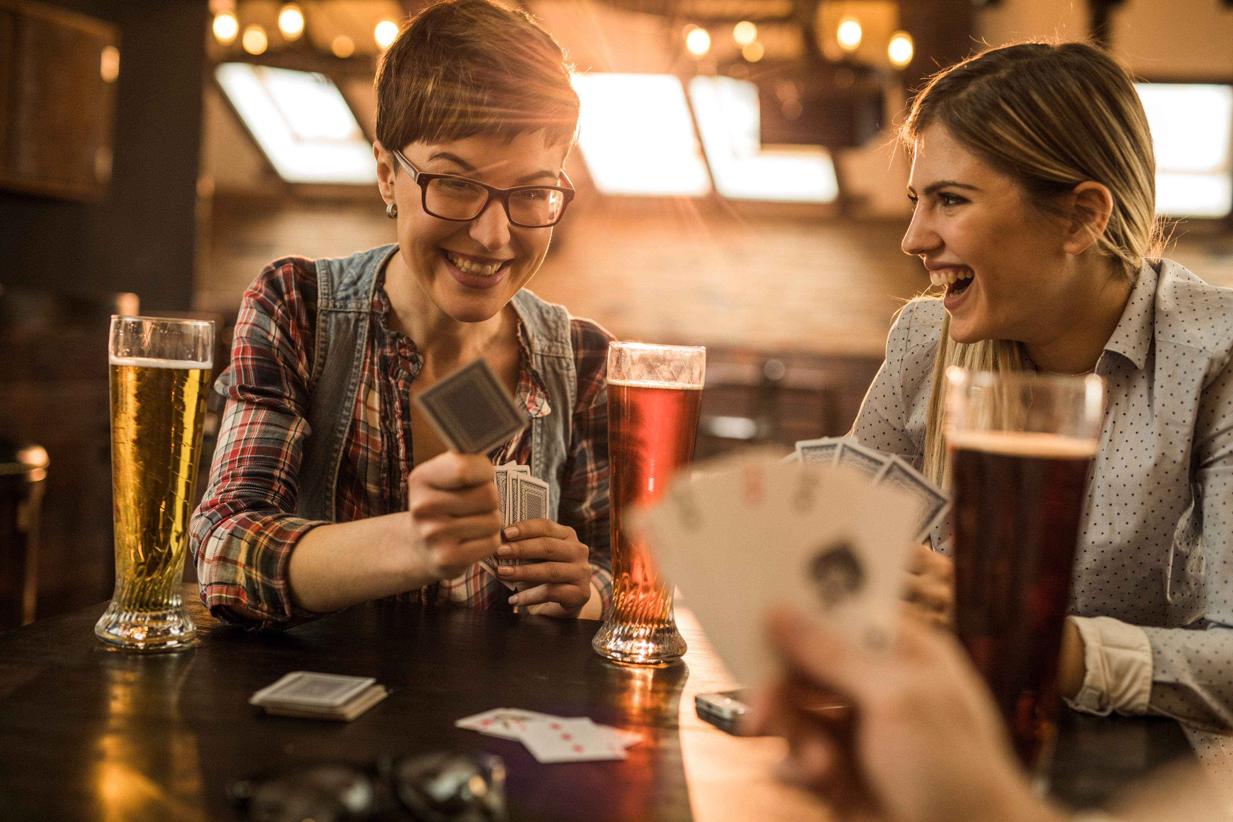 Two women smiling and laughing while playing cards and drinking beer at a bar