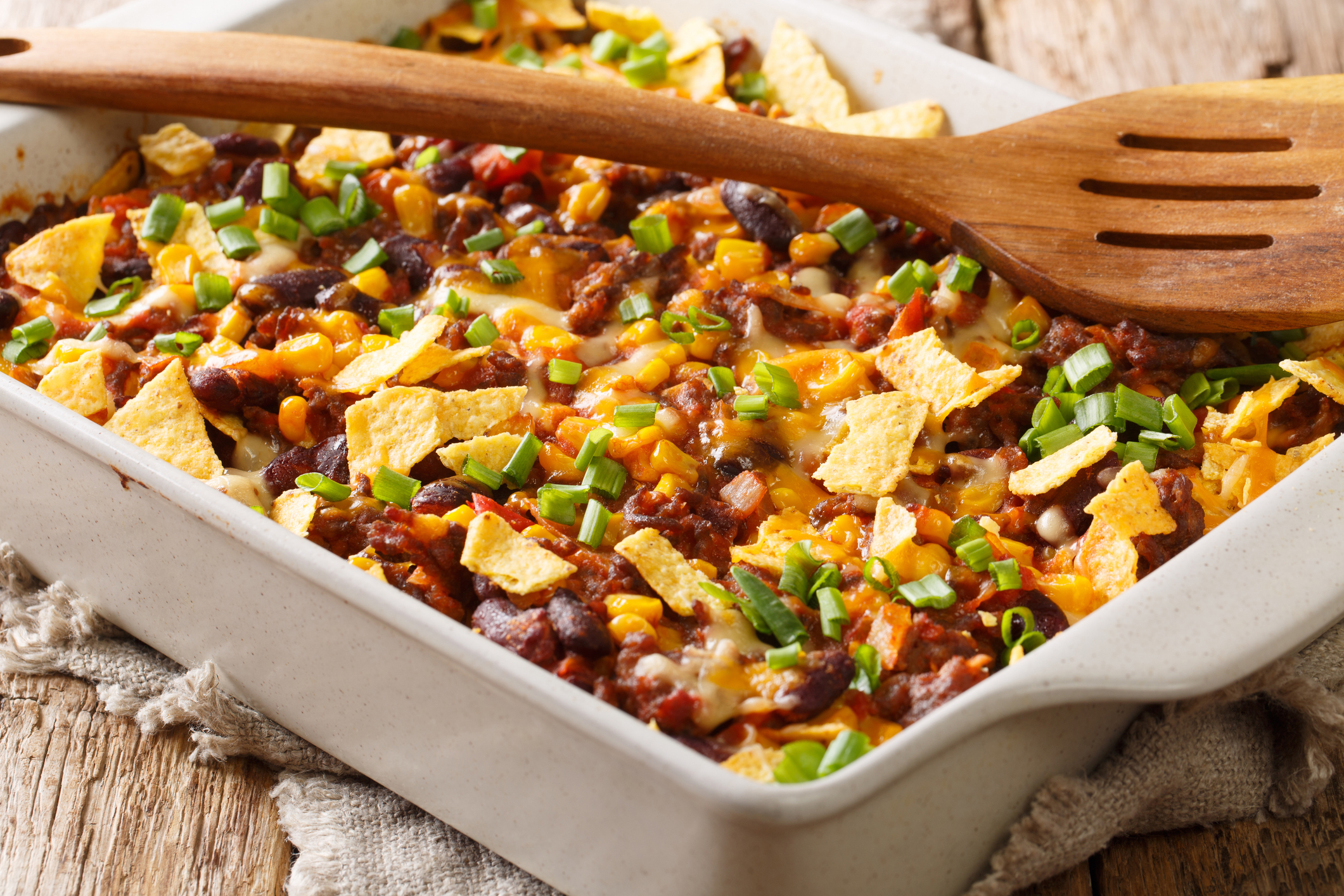 A close-up of a baked Mexican casserole dish topped with melted cheese, ground beef, beans, tortilla chips, and chopped green onions, with a wooden spoon on top