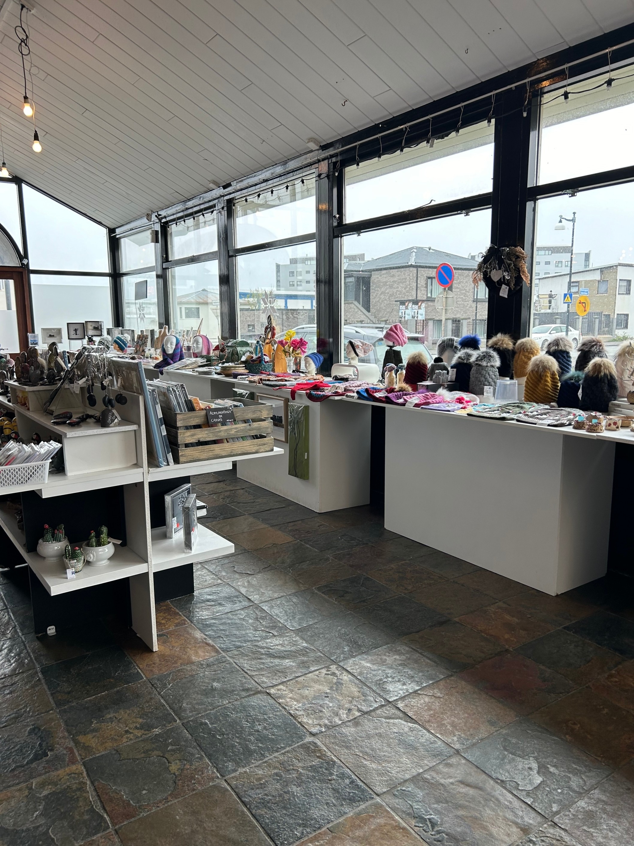 A bright, spacious shop with various items, including decorations and crafts, displayed on tables and shelves. Large windows illuminate the interior