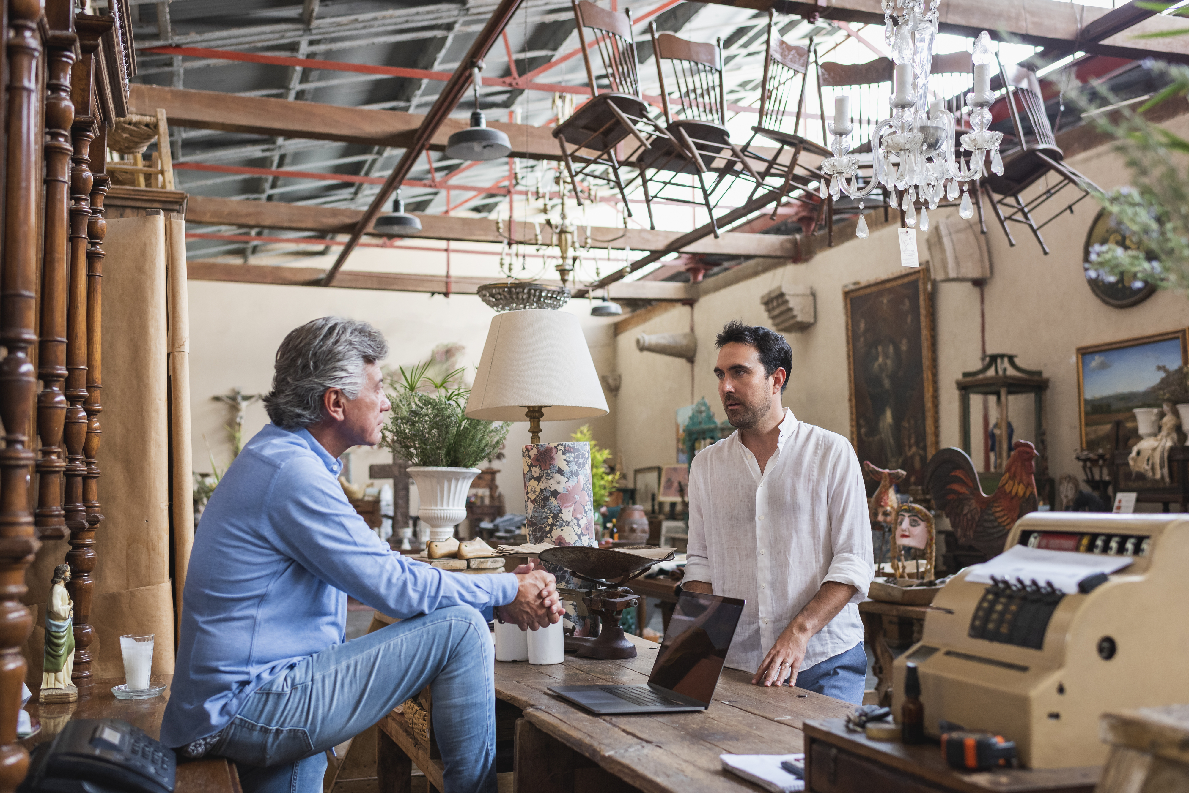 A man in a casual white shirt speaks to another man in a light blue shirt seated on a counter inside an antique shop filled with various vintage items