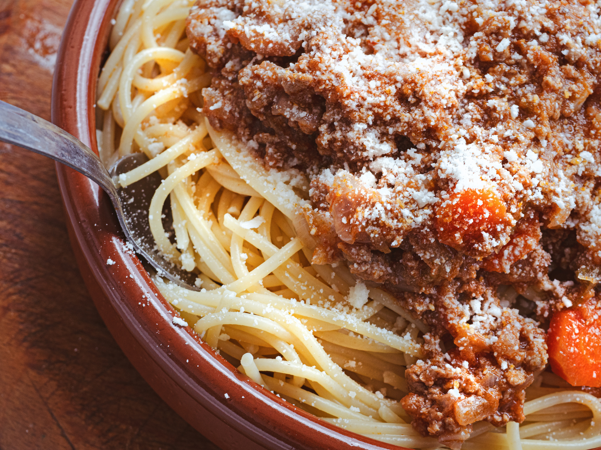 A bowl of spaghetti topped with a rich meat sauce and sprinkled with grated cheese. A fork is resting in the dish