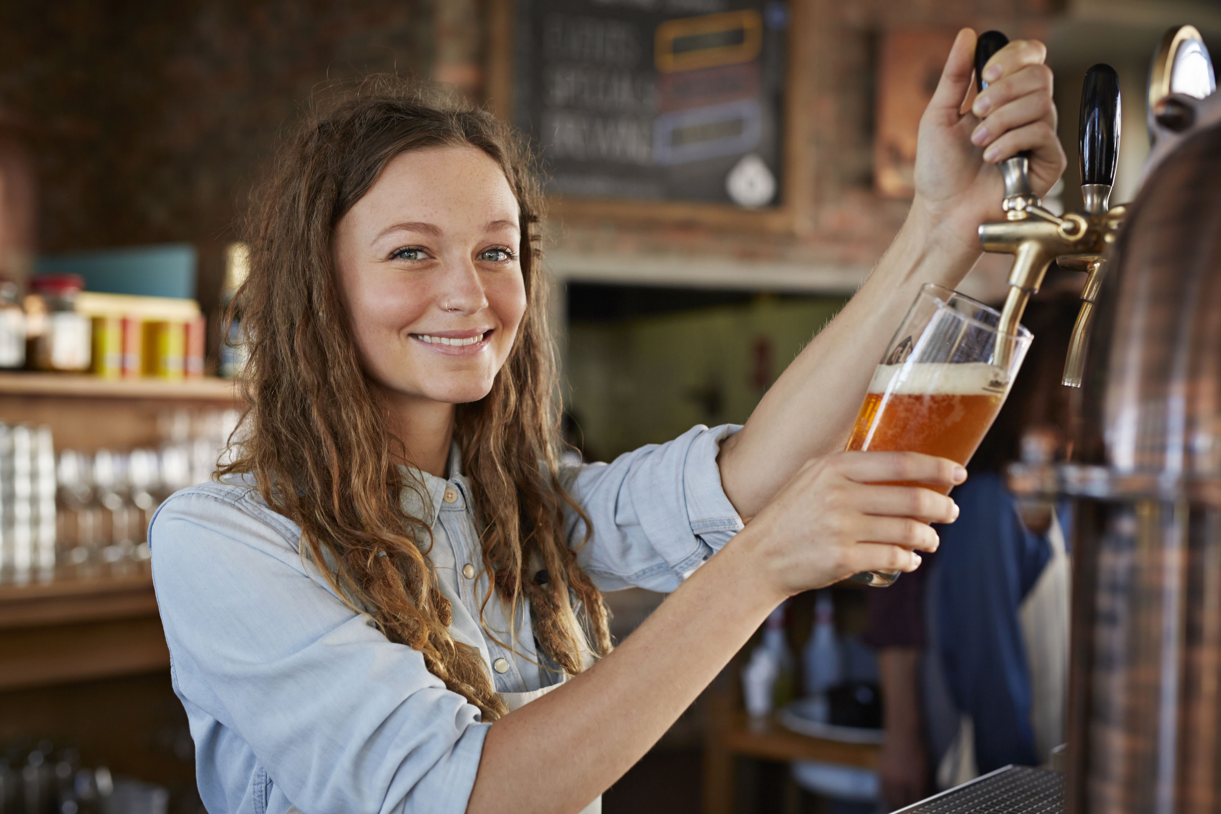 Smiling woman in casual attire pours a beer from a tap in a bar setting. Background shows bar decor and people socializing