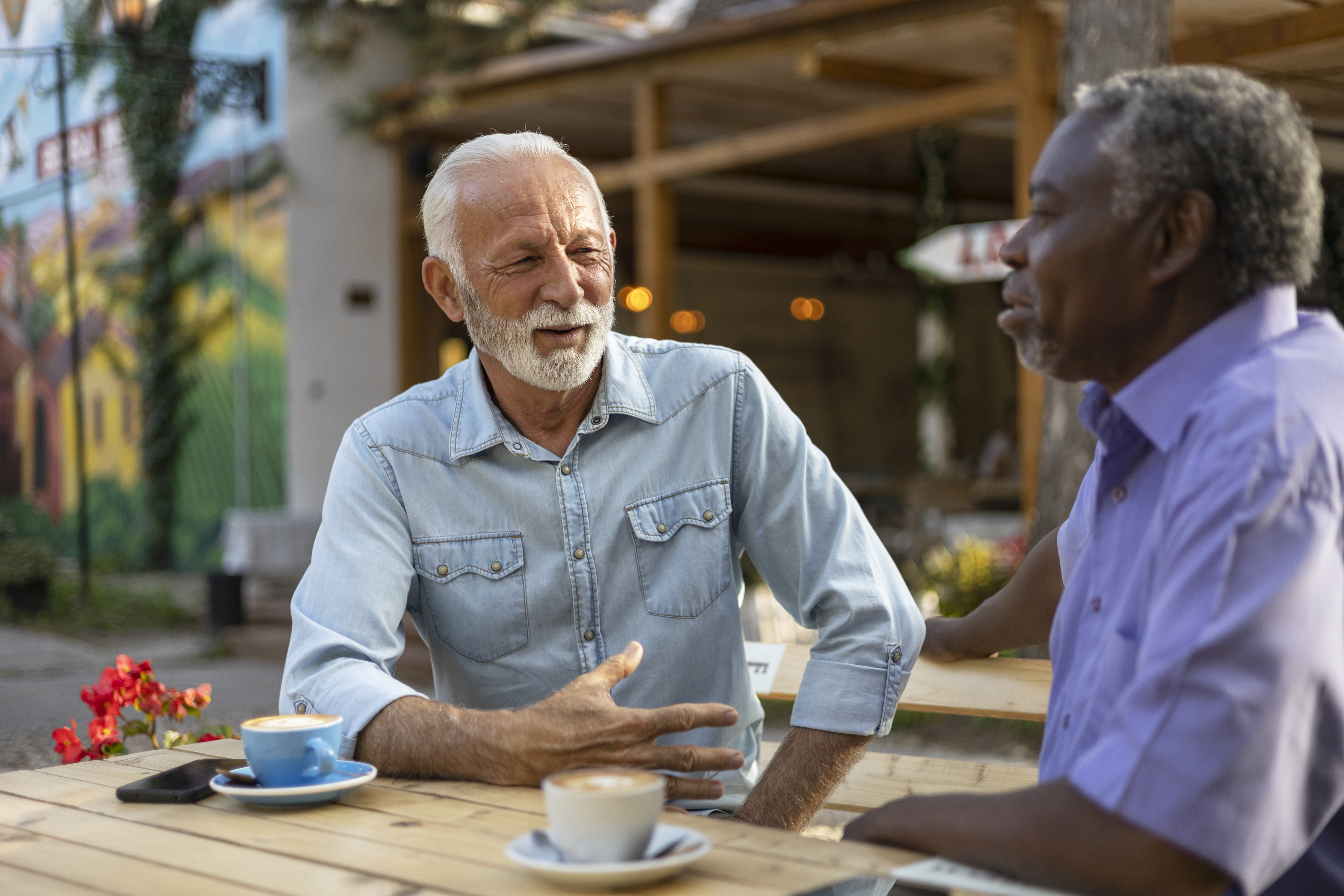 Two men, one in a light blue button-up shirt and the other in a purple short-sleeve shirt, converse at an outdoor café with coffee cups on the table