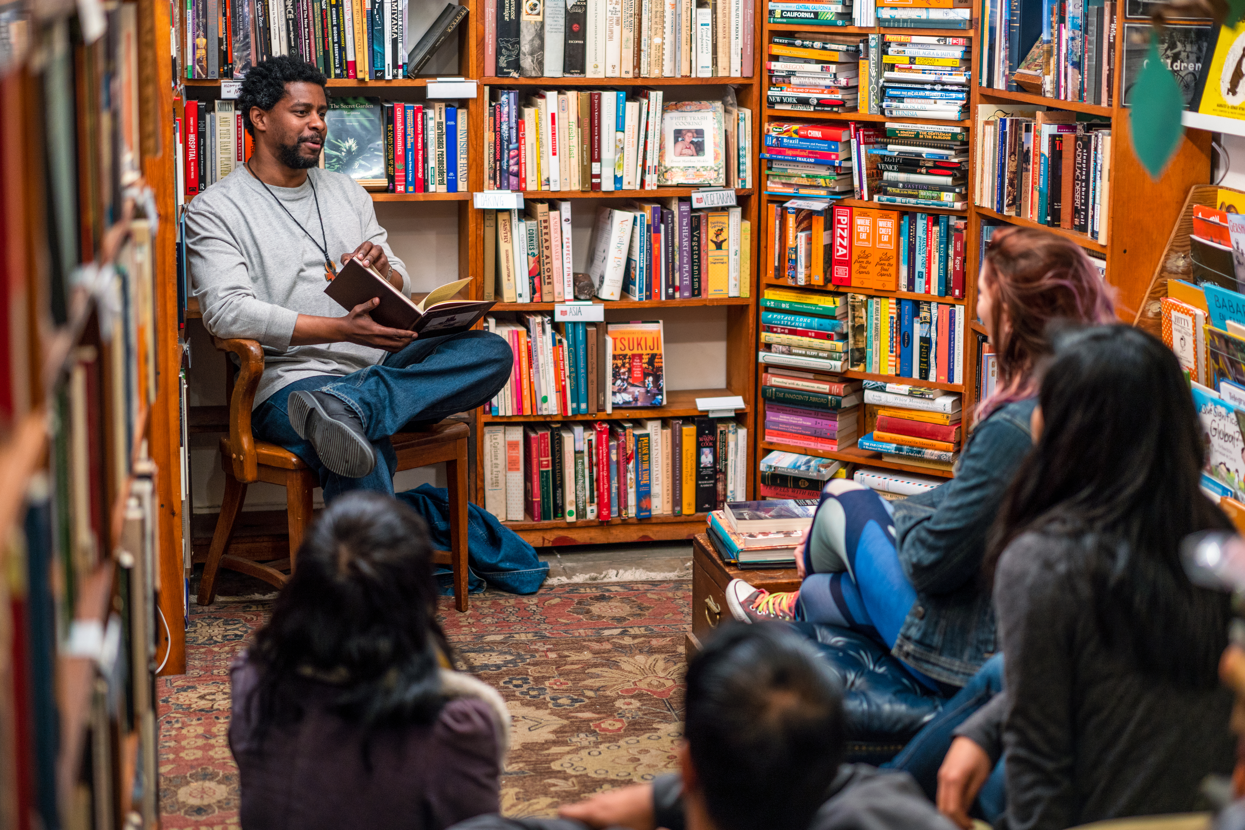 A person seated in a bookstore reads a book aloud to a small, seated audience. Shelves filled with books surround them