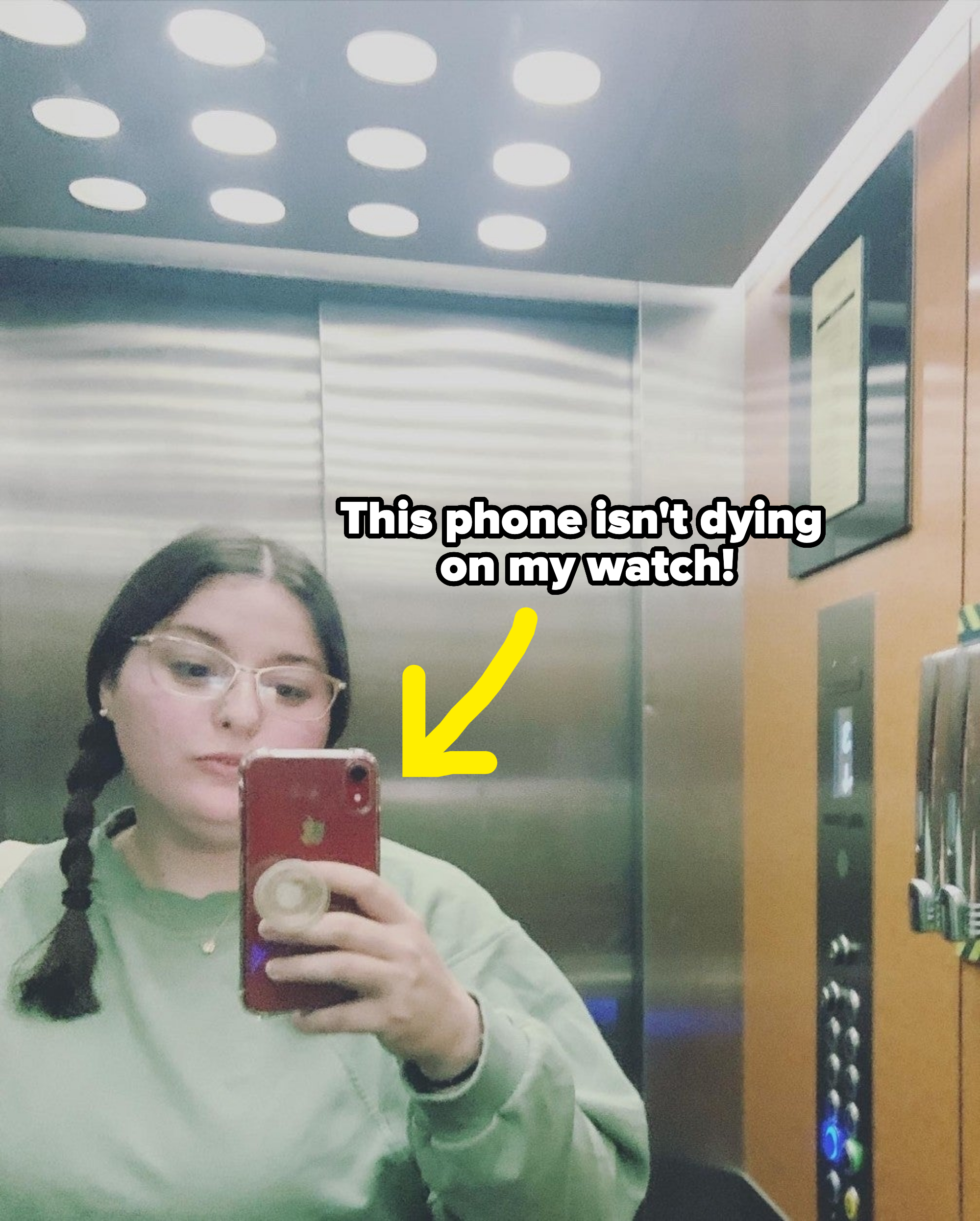 Kristen with glasses, wearing a sweater, takes a selfie in an elevator with reflective walls