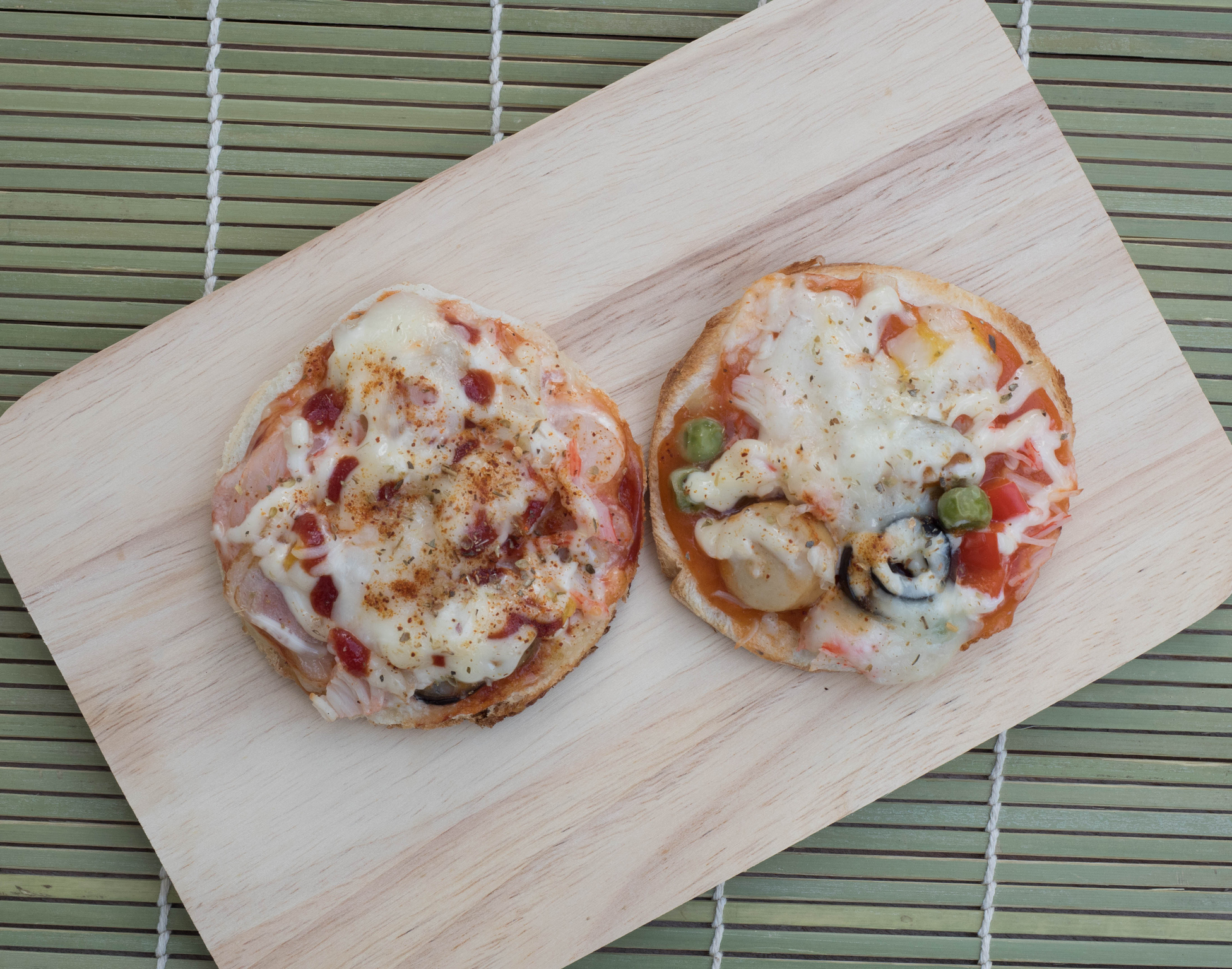 Two mini pizzas on a wooden board. Each pizza has a variety of toppings including cheese, vegetables, and possibly meat