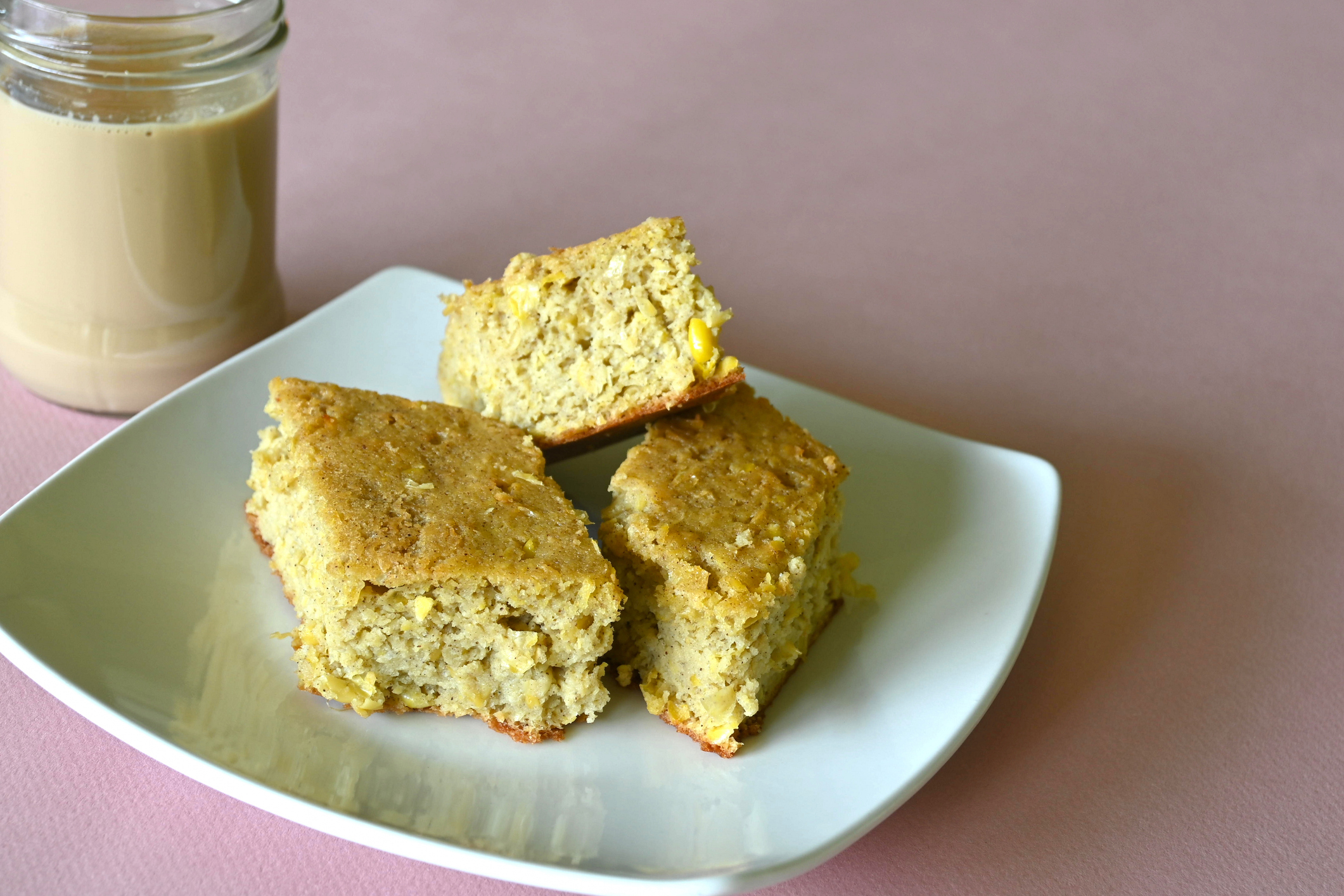 Three pieces of cornbread on a white plate, next to a jar of light-colored beverage, possibly coffee or tea, on a plain background