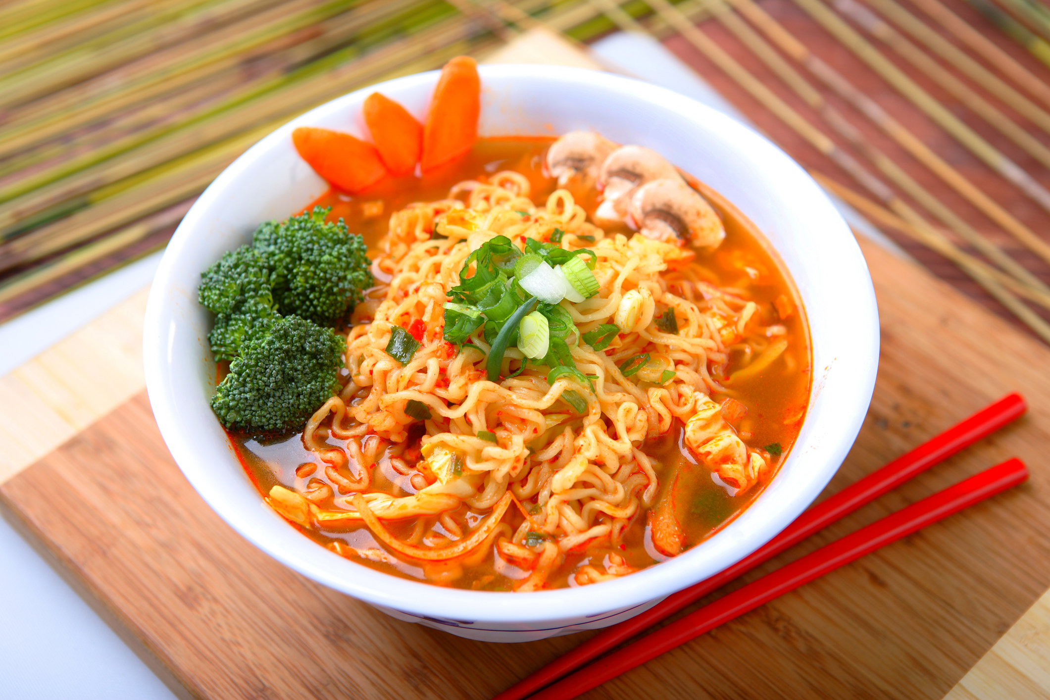Bowl of ramen noodles with broccoli, mushrooms, green onions, and carrot garnish on a wooden board with red chopsticks