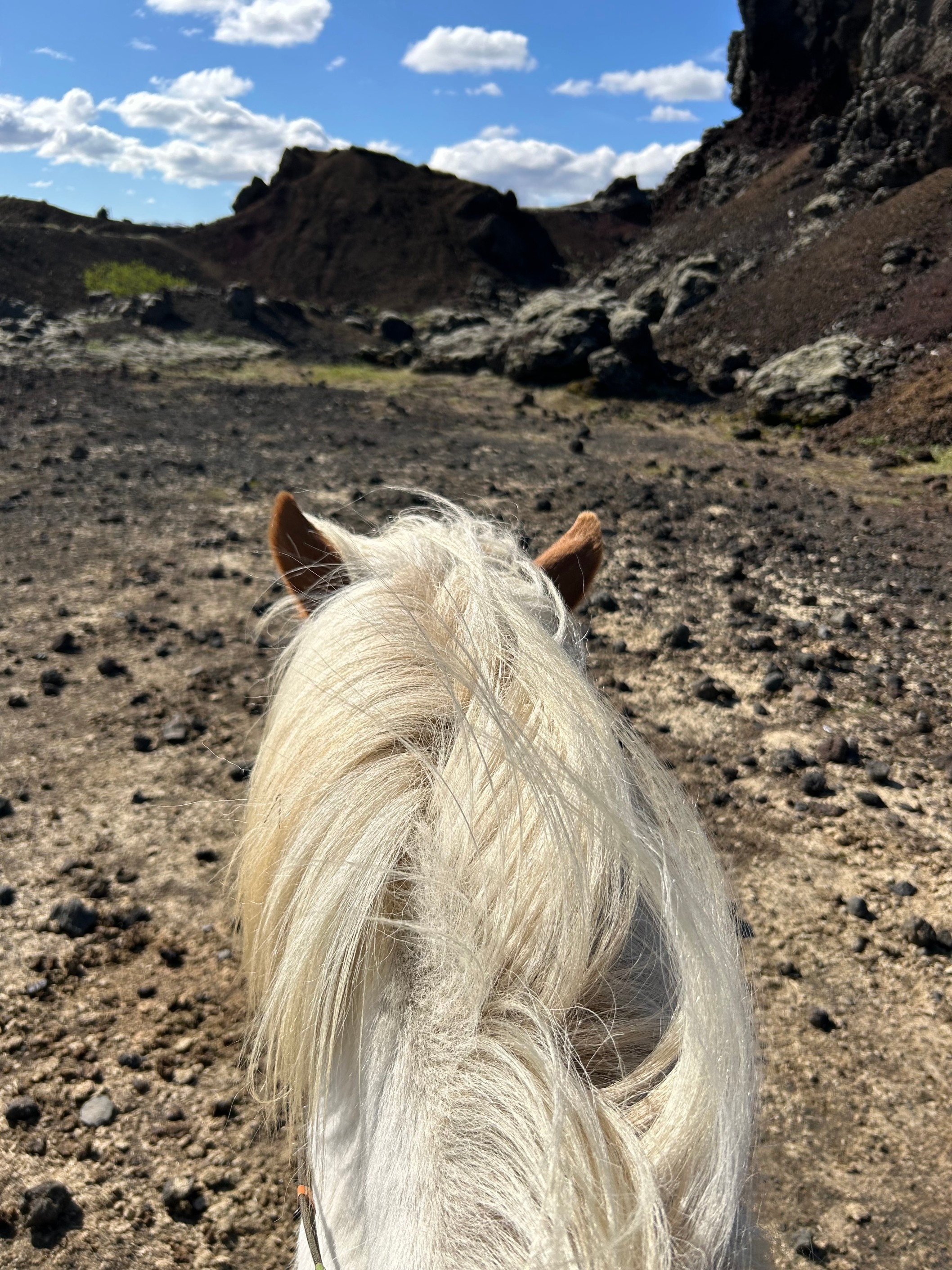 View from the rider's perspective of a horse walking on rocky terrain with hills in the background under a partly cloudy sky