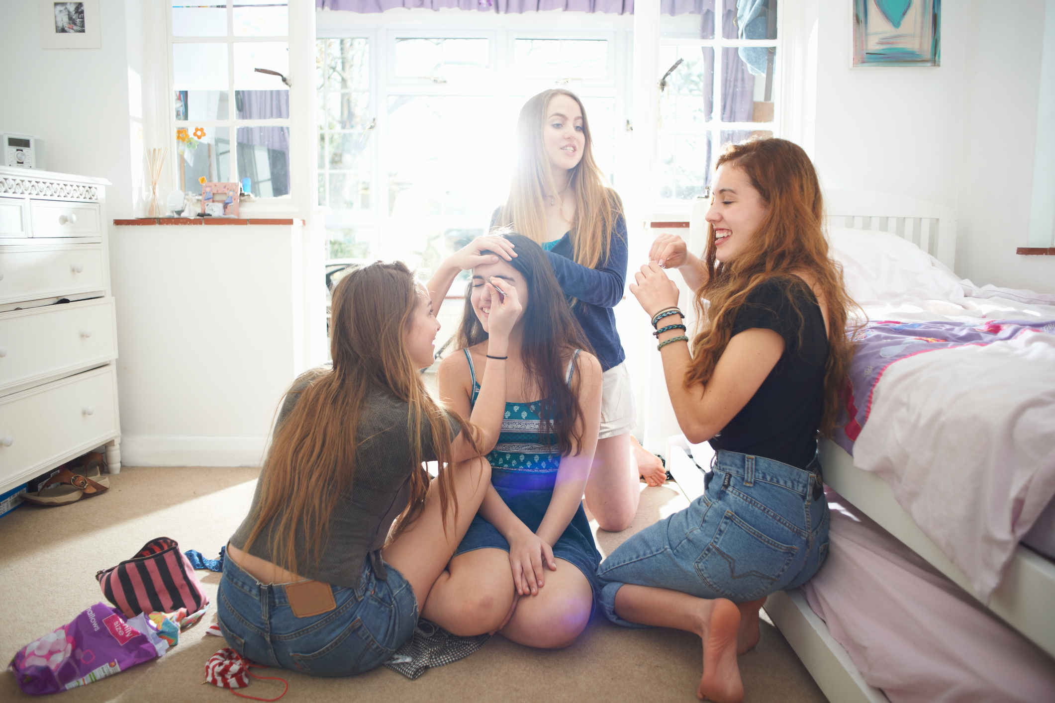 Four women are sitting on the floor of a bedroom, laughing and applying makeup on each other