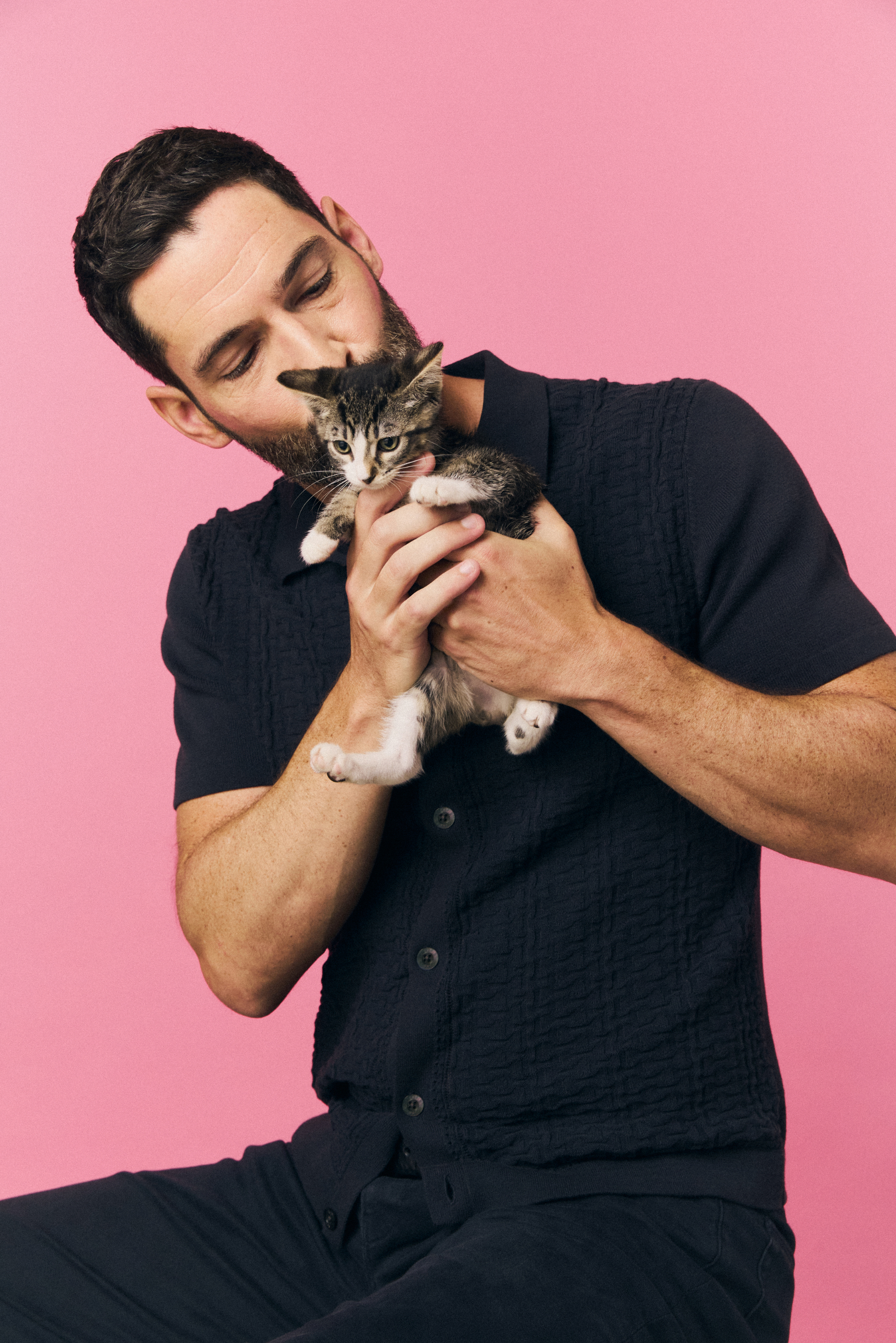 Tom Ellis holds a small kitten affectionately, posing against a pink background. He is dressed in a casual, textured short-sleeve shirt