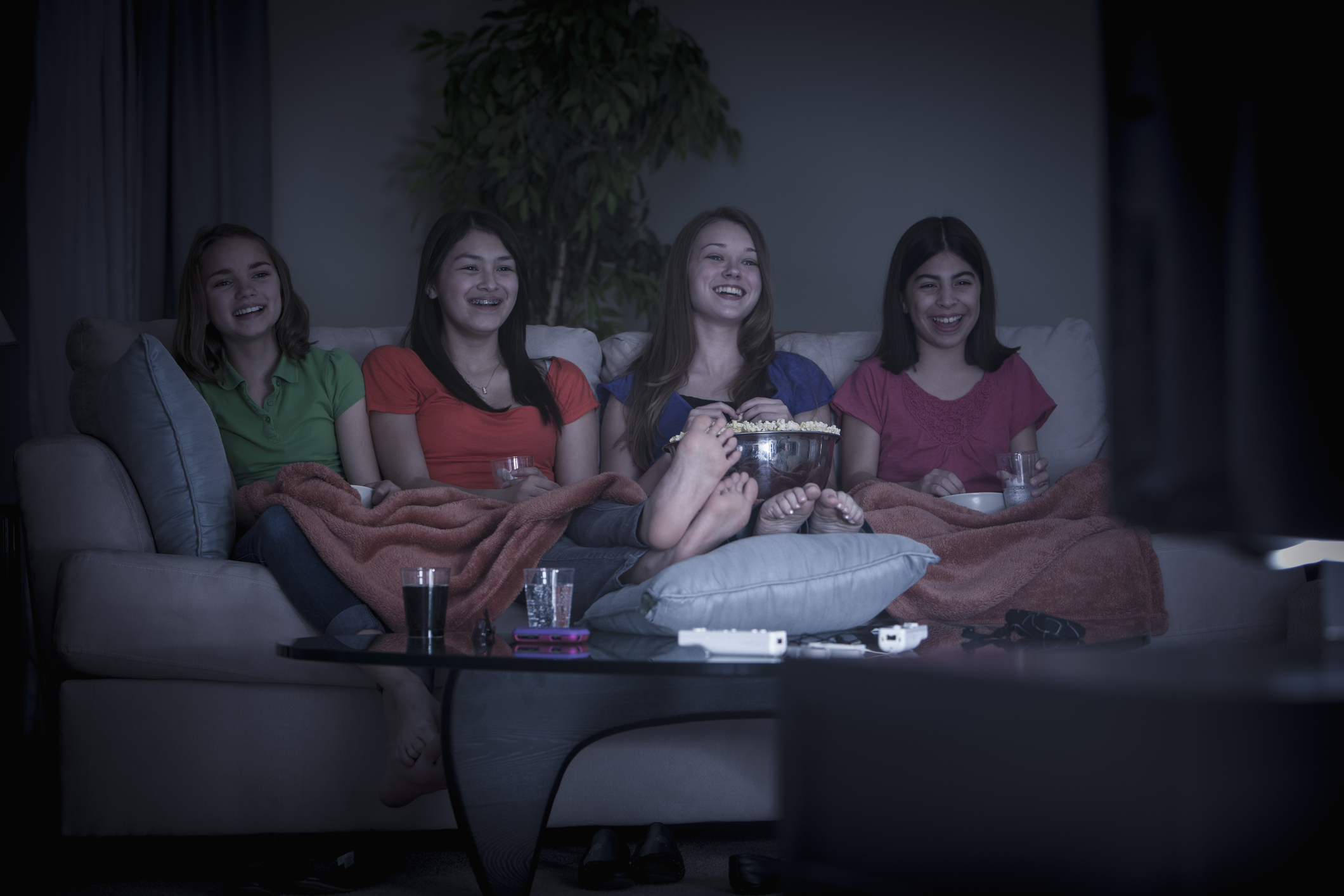 Four women, names unknown, sit on a couch under blankets, eating popcorn and watching TV. Two hold drinks, and all appear to be smiling and enjoying themselves