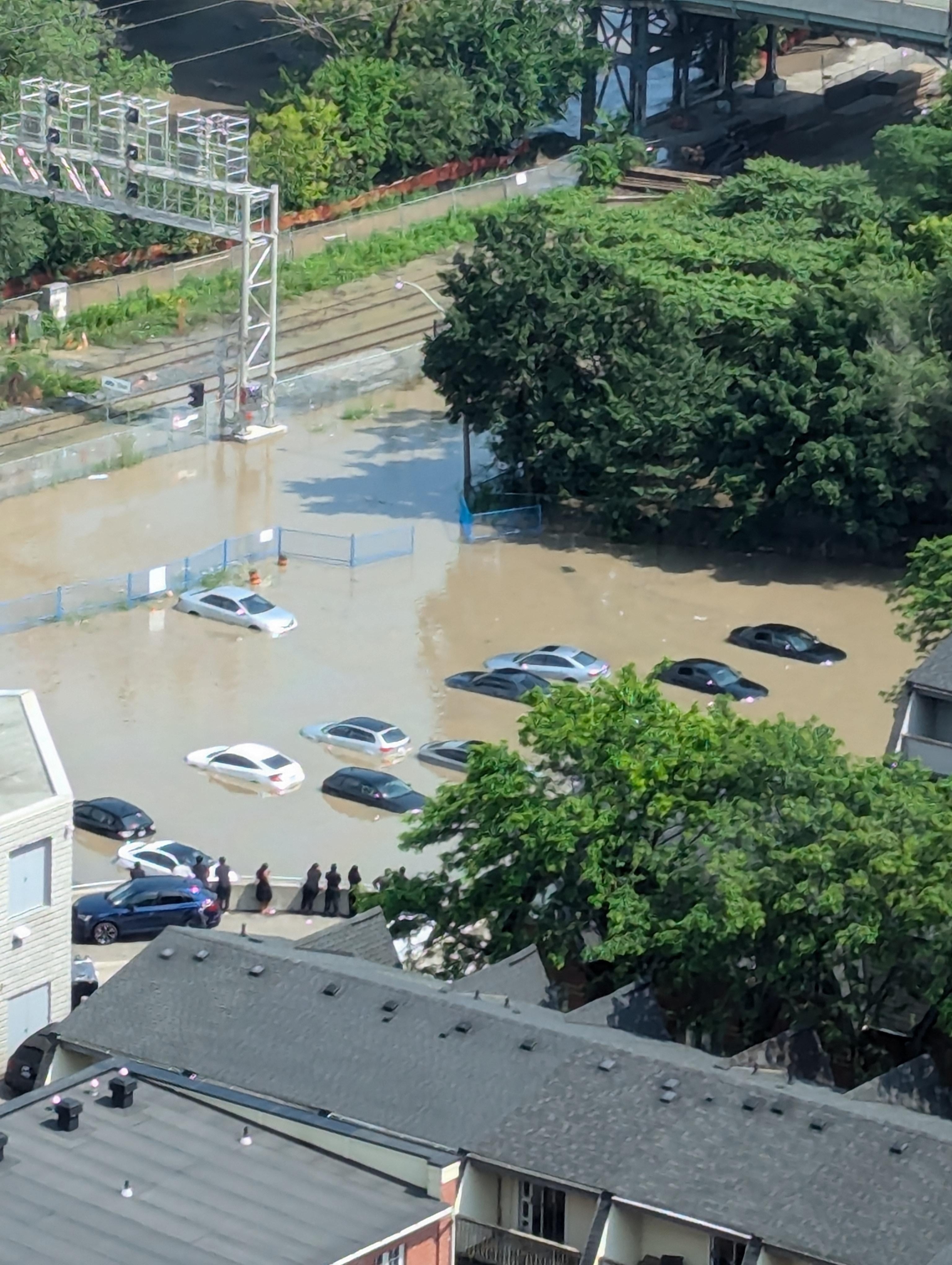 Several cars are submerged in floodwater near a railway track, surrounded by trees and residential buildings. People can be seen observing the scene