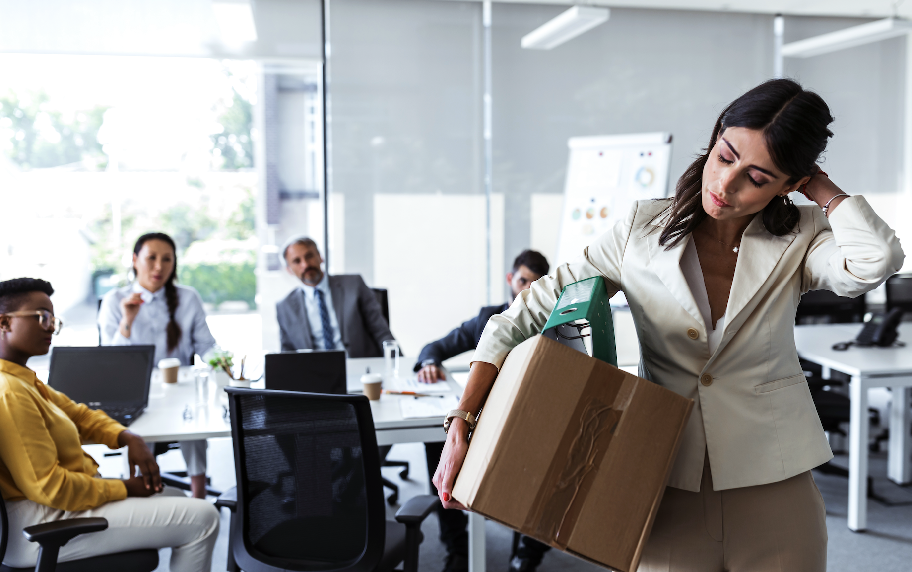 Woman in business attire carrying a box of belongings, indicating she's leaving her job, while four colleagues in business attire watch from a conference table