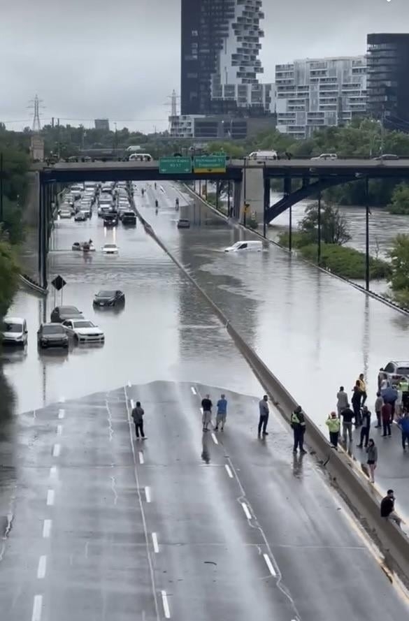 Flooded highway with partially submerged cars under an overpass, people standing on the median looking at the scene, and city buildings in the background