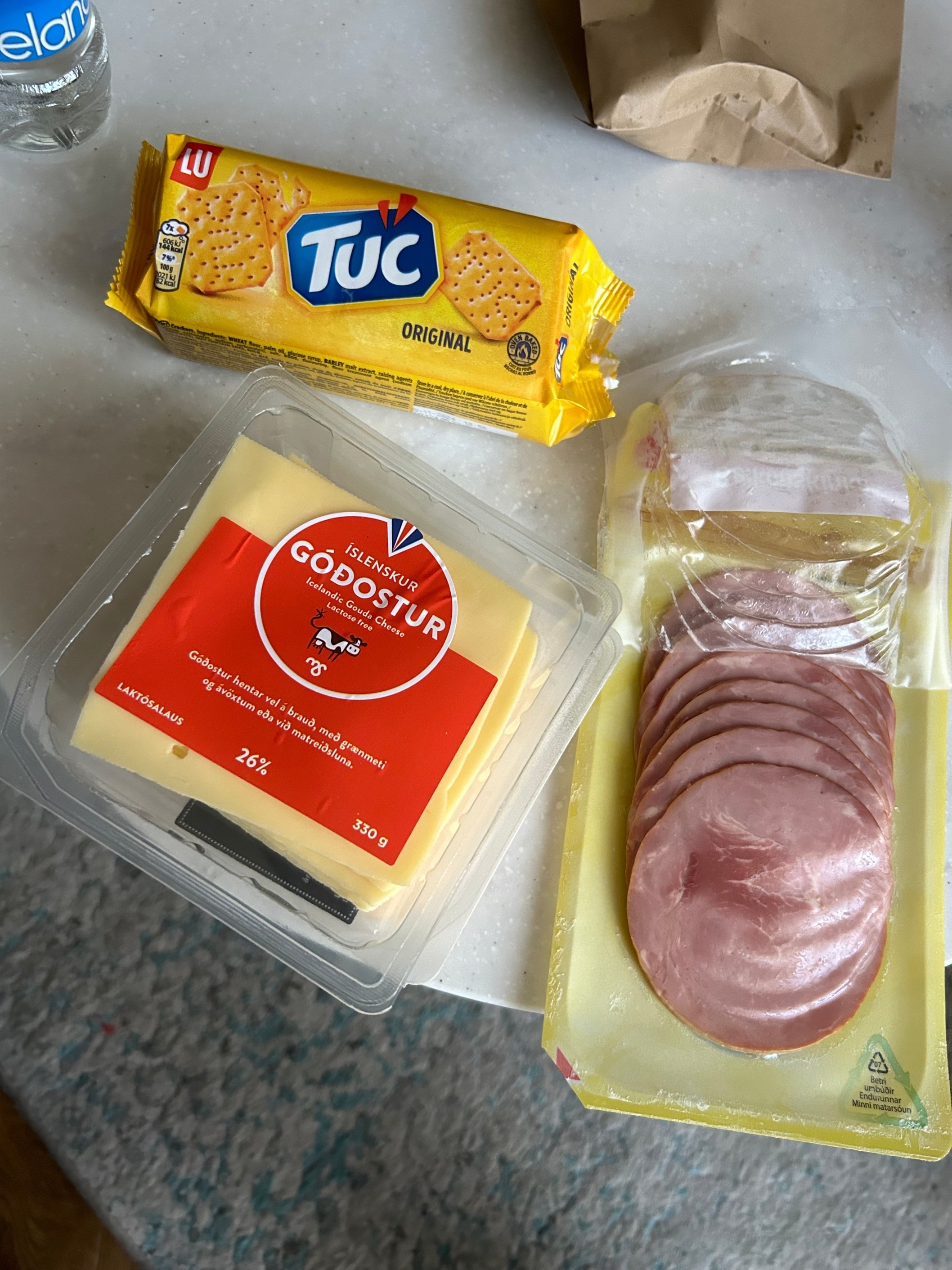 A pack of Tuc crackers, a container of sliced cheese labeled "Gouda," and a package of sliced ham are placed on a table