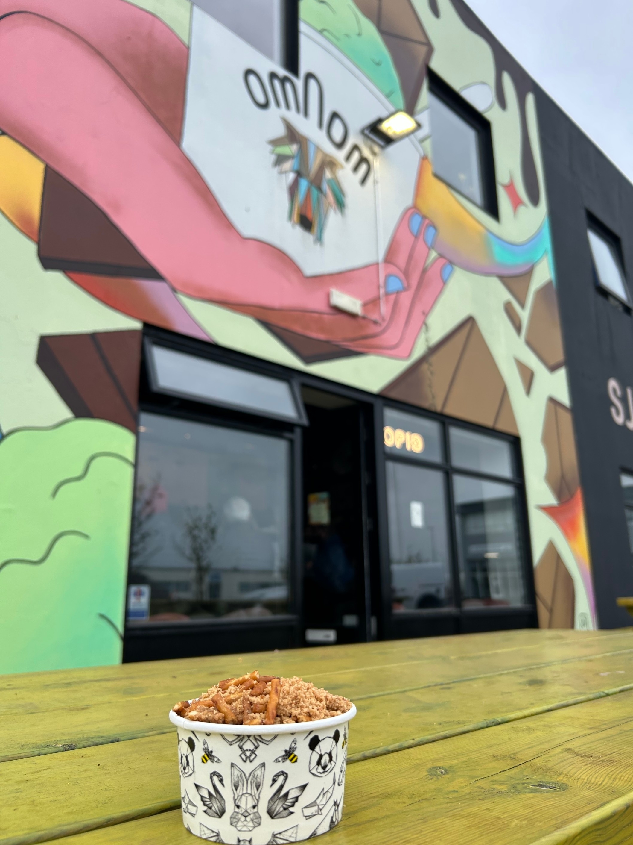 A colorful mural on a building exterior with "omnom" written at the top. In the foreground, a cup of food is on a wooden table