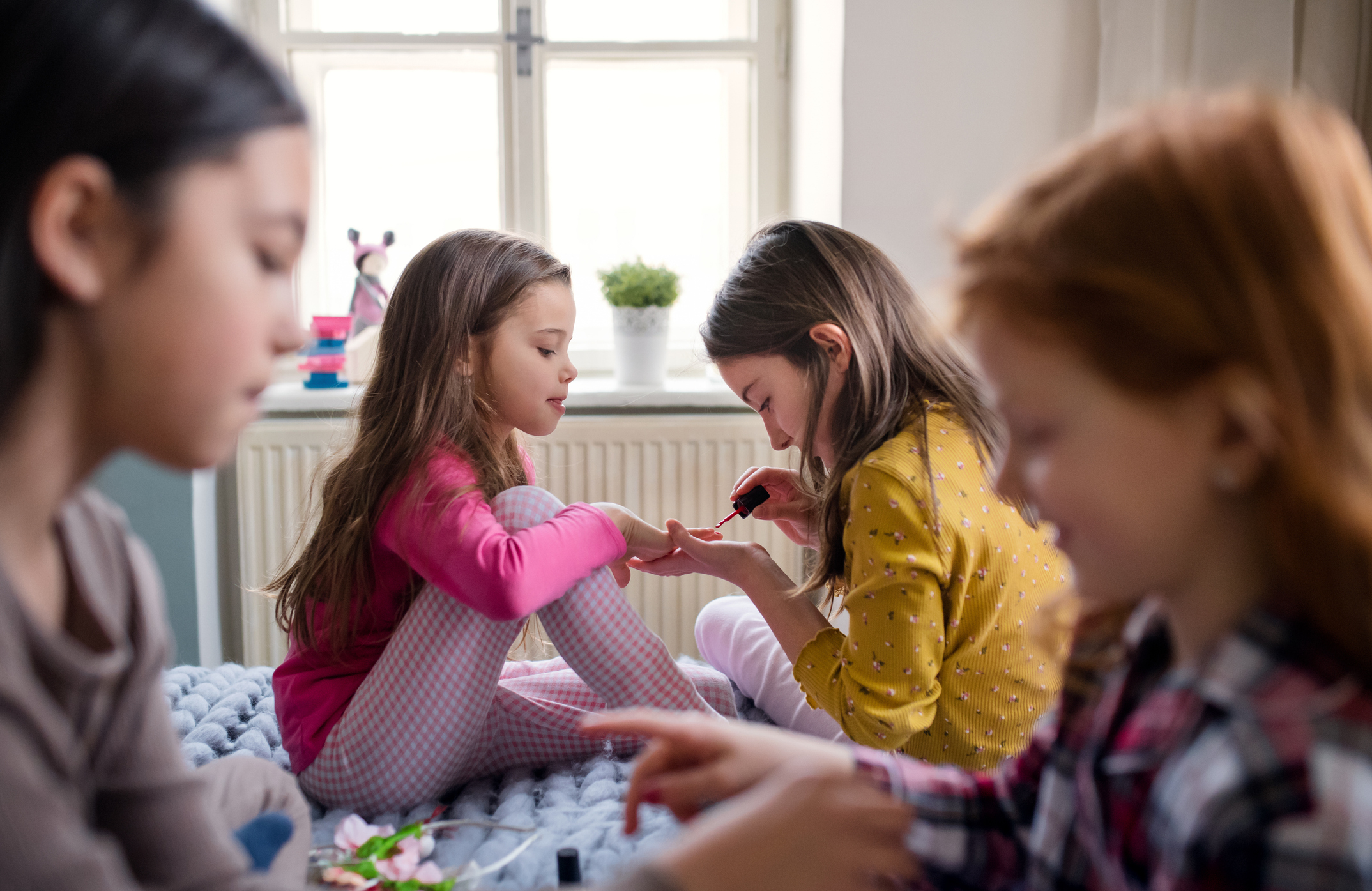 Four young girls, including two shown painting nails, sit together on a bed engaging in a fun activity
