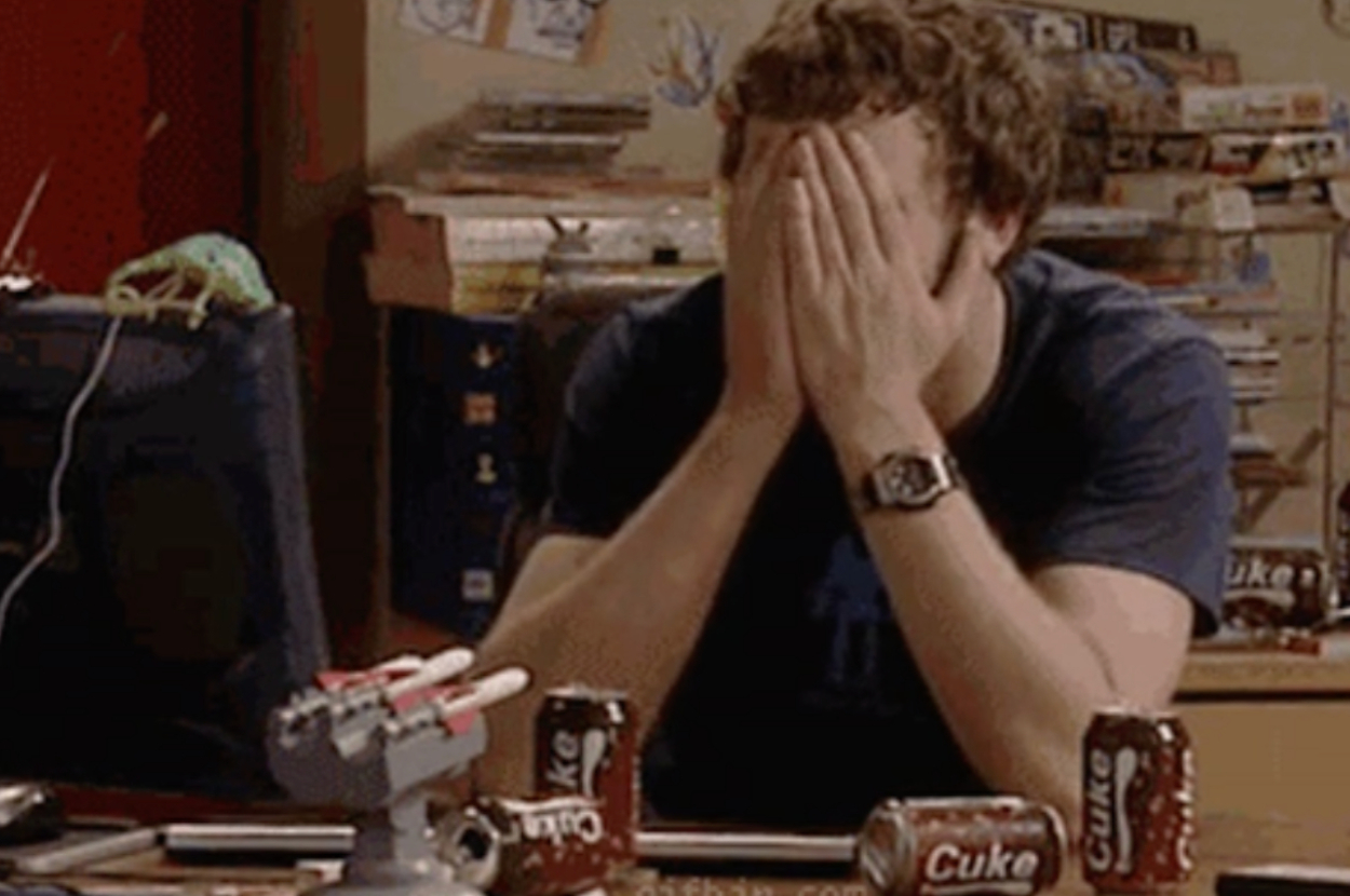 A person sits at a cluttered desk, with hands covering their face, surrounded by Coca-Cola cans and various objects, indicating frustration or stress