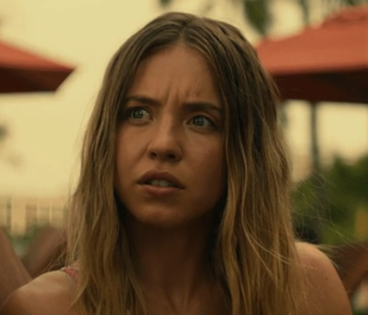 Sydney Sweeney with long, wavy hair, looks surprised or confused, outdoors with blurred umbrellas and greenery in the background