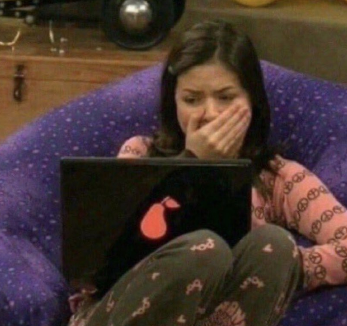 Miranda Cosgrove, sitting on a bean bag, wearing patterned pajamas, covering her mouth in shock while looking at a laptop