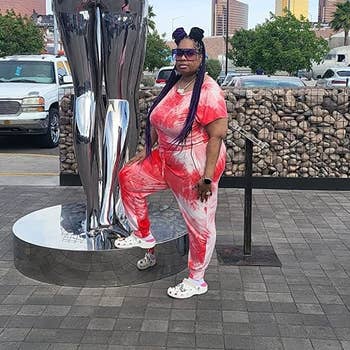 Person with agelong braids, wearing a vibrant tie-dye outfit and achromatic sandals, posing adjacent to a metallic creation sculpture successful an outdoor country with buildings and thenar trees