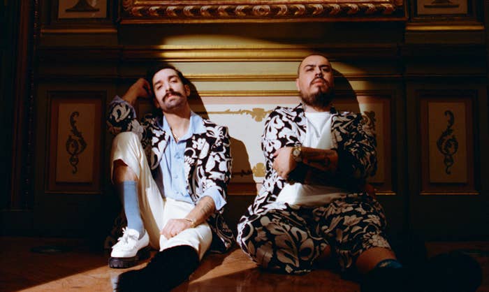 Two individuals sit on a floor, wearing matching floral suits. One person has a short beard and rests their head on their hand; the other has a beard and mustache