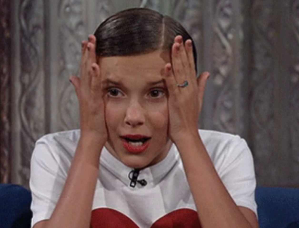 Millie Bobby Brown holds her face with both hands, looking surprised while wearing a white shirt with a red heart