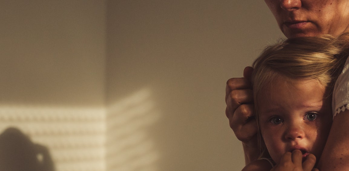 A woman wearing a white blouse holds a young child, who appears worried. They are in a dimly lit room with shadows cast on the walls
